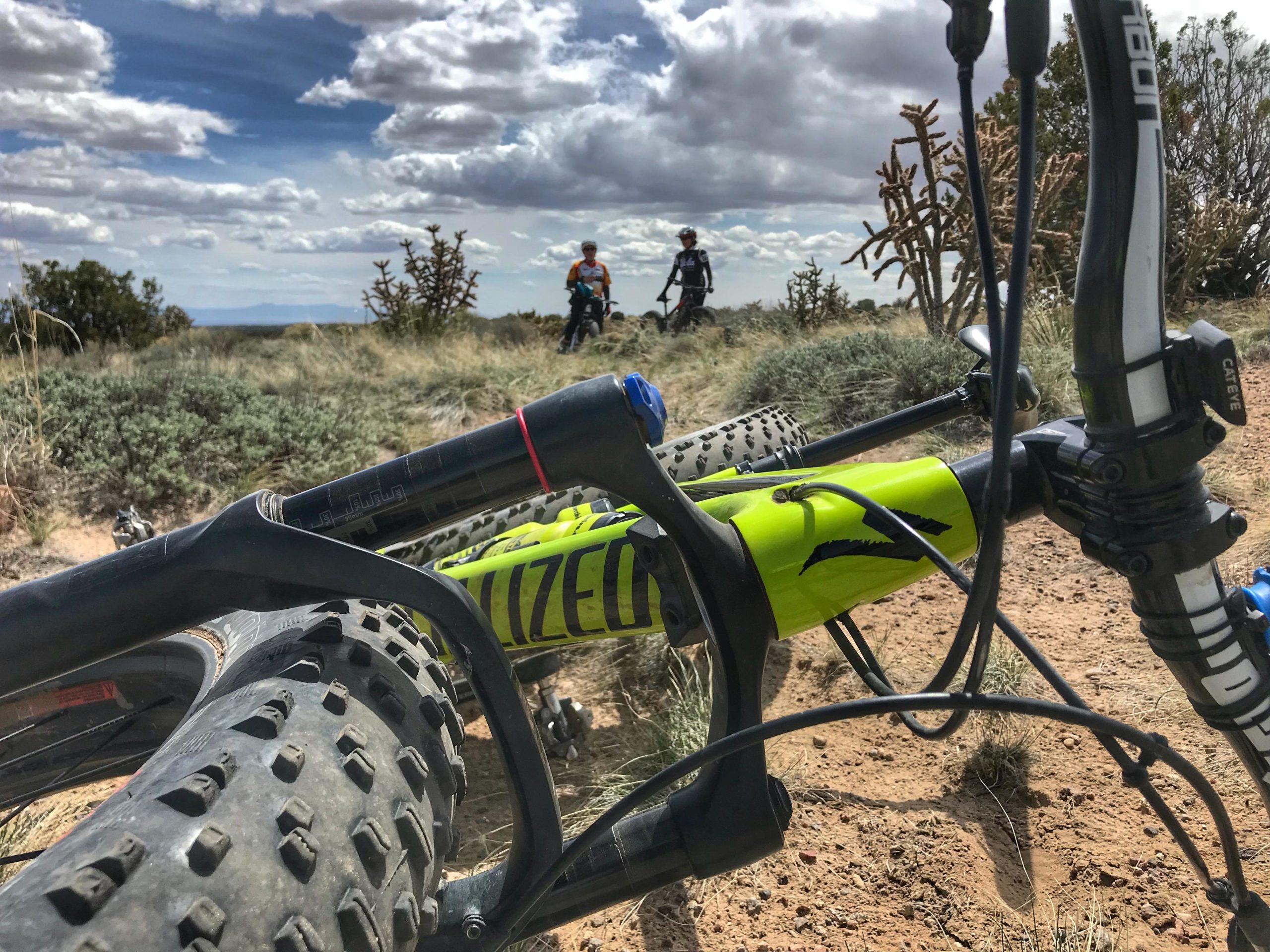 A close-up view of a mountain bike in the foreground, showcasing its large tires and vibrant yellow frame. In the background, two cyclists are visible riding on a trail surrounded by desert vegetation and a partly cloudy sky. The landscape features dry grass and shrubs, creating a scenic outdoor environment. Mariposa Fat Bike Trails mountain bike trail.