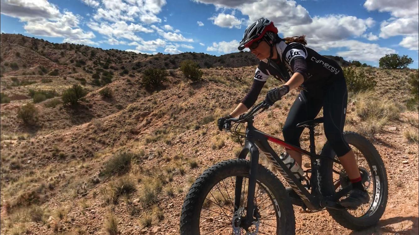 A cyclist riding a fat bike on a rocky, arid trail surrounded by hills and sparse vegetation under a partly cloudy sky. The rider is wearing a helmet and cycling gear, focused on navigating the terrain. Mariposa Fat Bike Trails mountain bike trail.