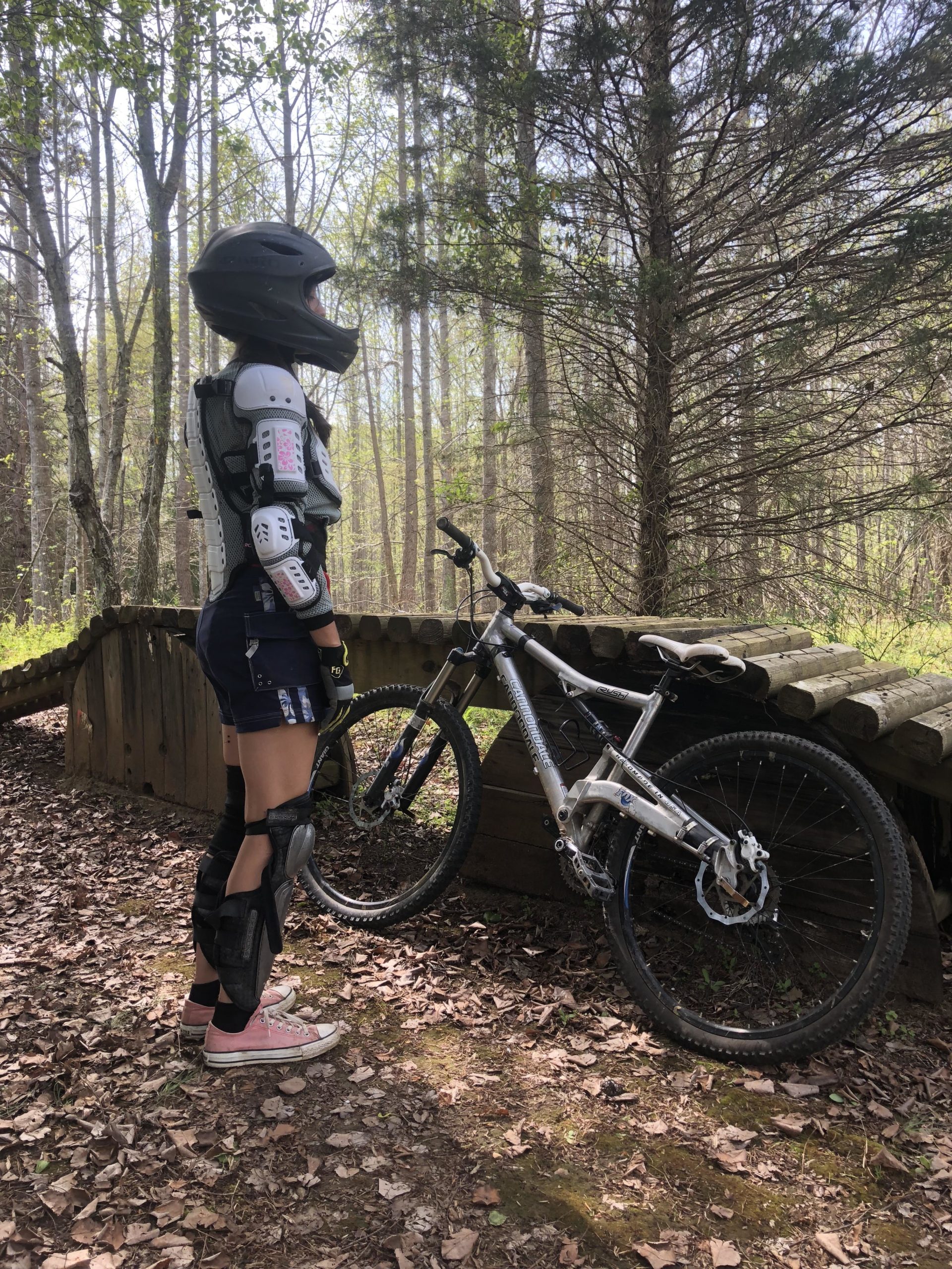 A person wearing a black helmet and protective gear is standing next to a mountain bike. They are positioned on a forest trail surrounded by trees and fallen leaves, looking off into the distance. The bike is silver and leans against a wooden structure. Angler's Ridge mountain bike trail.