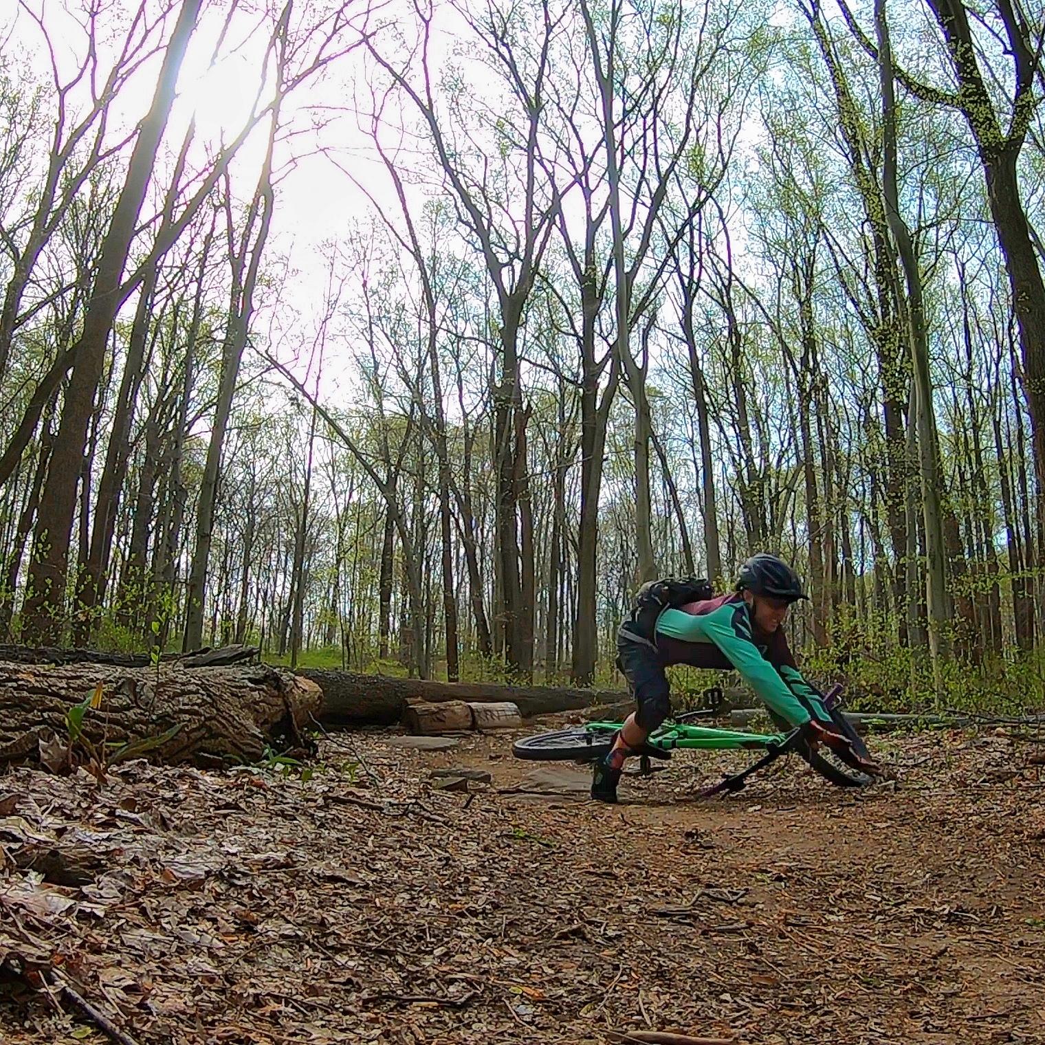A mountain biker leans low over their bike as they navigate a dirt trail in a wooded area, surrounded by tall trees and logs on the ground. The sun is shining through the branches, illuminating the scene. Brandywine State Park mountain bike trail.