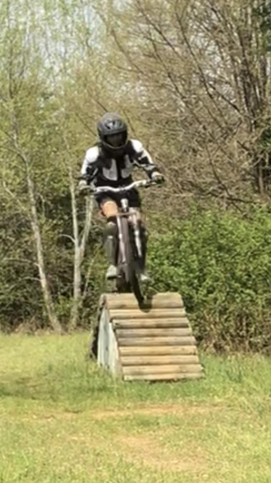 A person wearing a black helmet and protective gear is mid-air while riding a mountain bike over a wooden ramp in a grassy area surrounded by trees. Angler's Ridge mountain bike trail.