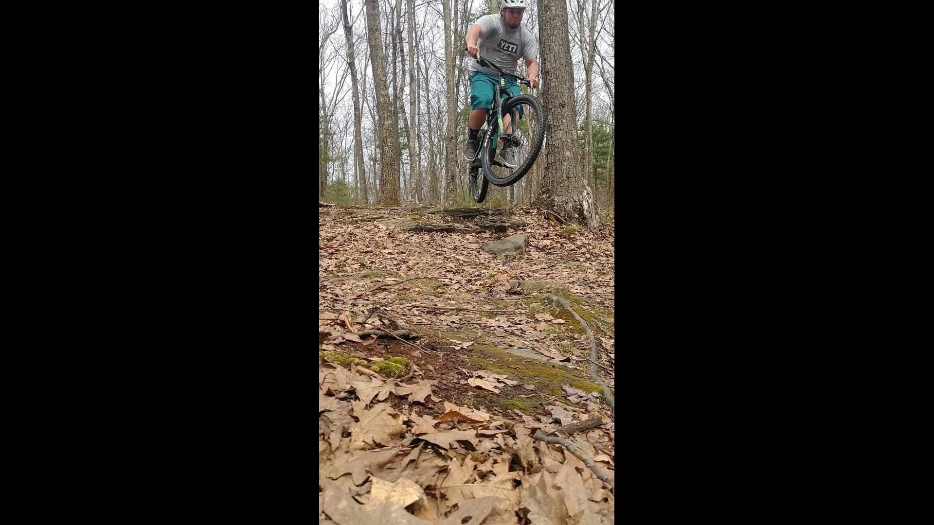 A mountain biker performing a jump over a rocky terrain, surrounded by trees and covered in fallen leaves. The cyclist is wearing a helmet and sportswear, showcasing action and excitement in a natural outdoor setting. Moon Lake Park mountain bike trail.
