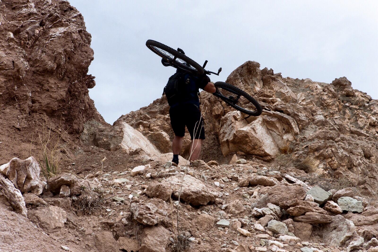 A cyclist carrying a mountain bike over rocky terrain while climbing a steep, uneven hillside. The landscape features various sizes of rocks and earthy colors, under a cloudy sky. White Ridge Bike Trails mountain bike trail.