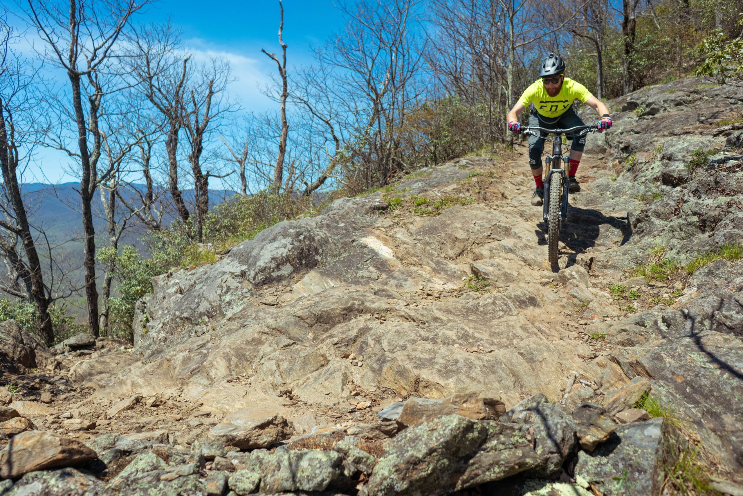 A mountain biker navigating a rocky trail with bare trees and blue skies in the background, showcasing a challenging outdoor biking environment. Pilot Rock mountain bike trail.