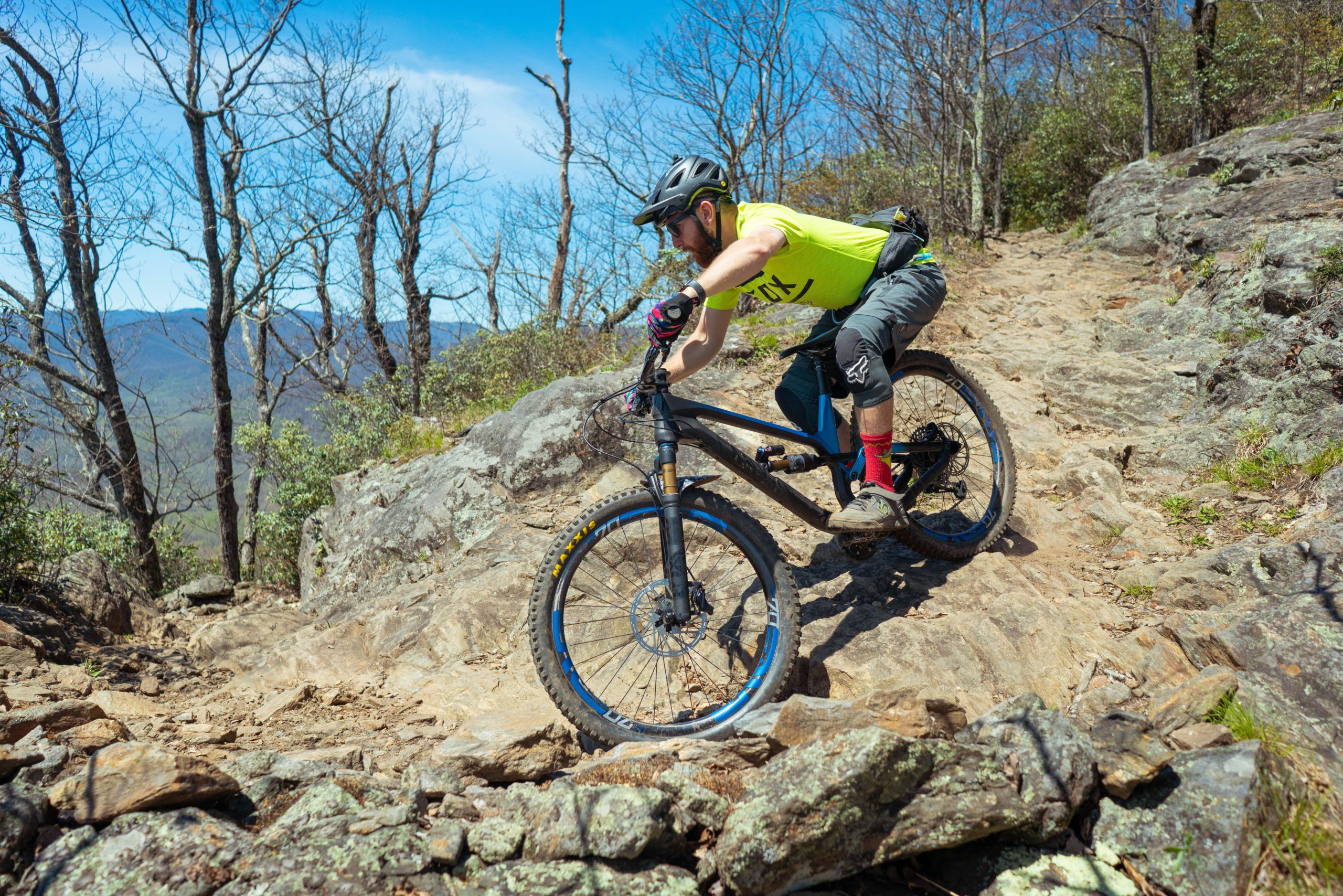 A mountain biker navigating a rocky trail, wearing a bright yellow shirt and gray shorts, with red socks and gloves. The background features a clear blue sky and bare trees, with distant mountains visible. The biker is focused, leaning forward as he rides over uneven terrain. Pilot Rock mountain bike trail.