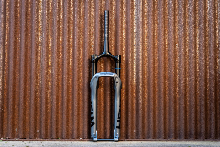 A bicycle fork leaning against a corrugated rust-colored metal wall. The fork features a dark black and light gray design, with the logo "Cane Creek" visible. The scene is simple and industrial in appearance, focusing on the bicycle component.