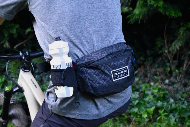 A person wearing a gray t-shirt is seen from the back, riding a bicycle. They have a black Dakine waist pack secured around their waist, which holds a white water bottle. The background features greenery and parts of the bike.