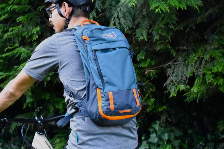 A person wearing a helmet and a gray shirt is riding a bicycle, showcasing a blue and orange backpack. The background features lush green foliage.