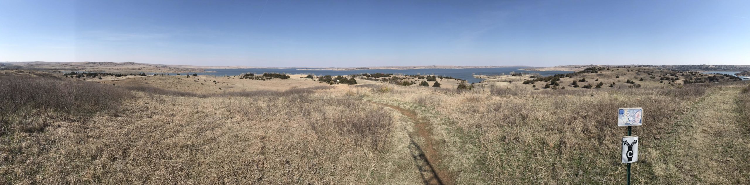 A panoramic view of a grassy landscape overlooking a lake, with rolling hills in the background and a clear blue sky. A trail sign is visible in the foreground, marking a walking path, while sparse trees dot the scenery. Switchgrass mountain bike trail.