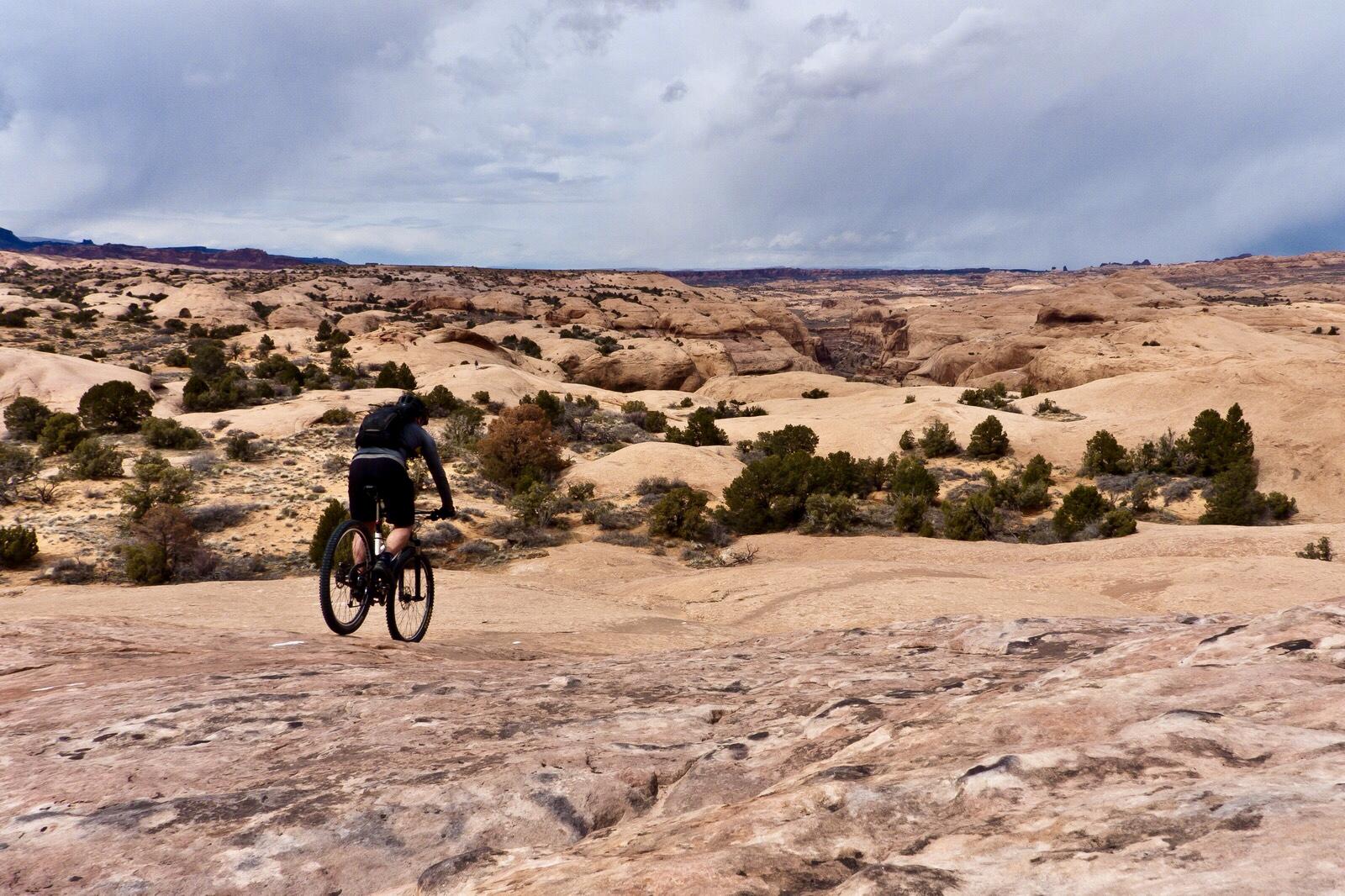 A mountain biker riding on a rocky terrain, surrounded by a vast landscape of natural rock formations and sparse vegetation under a cloudy sky. Slickrock mountain bike trail.