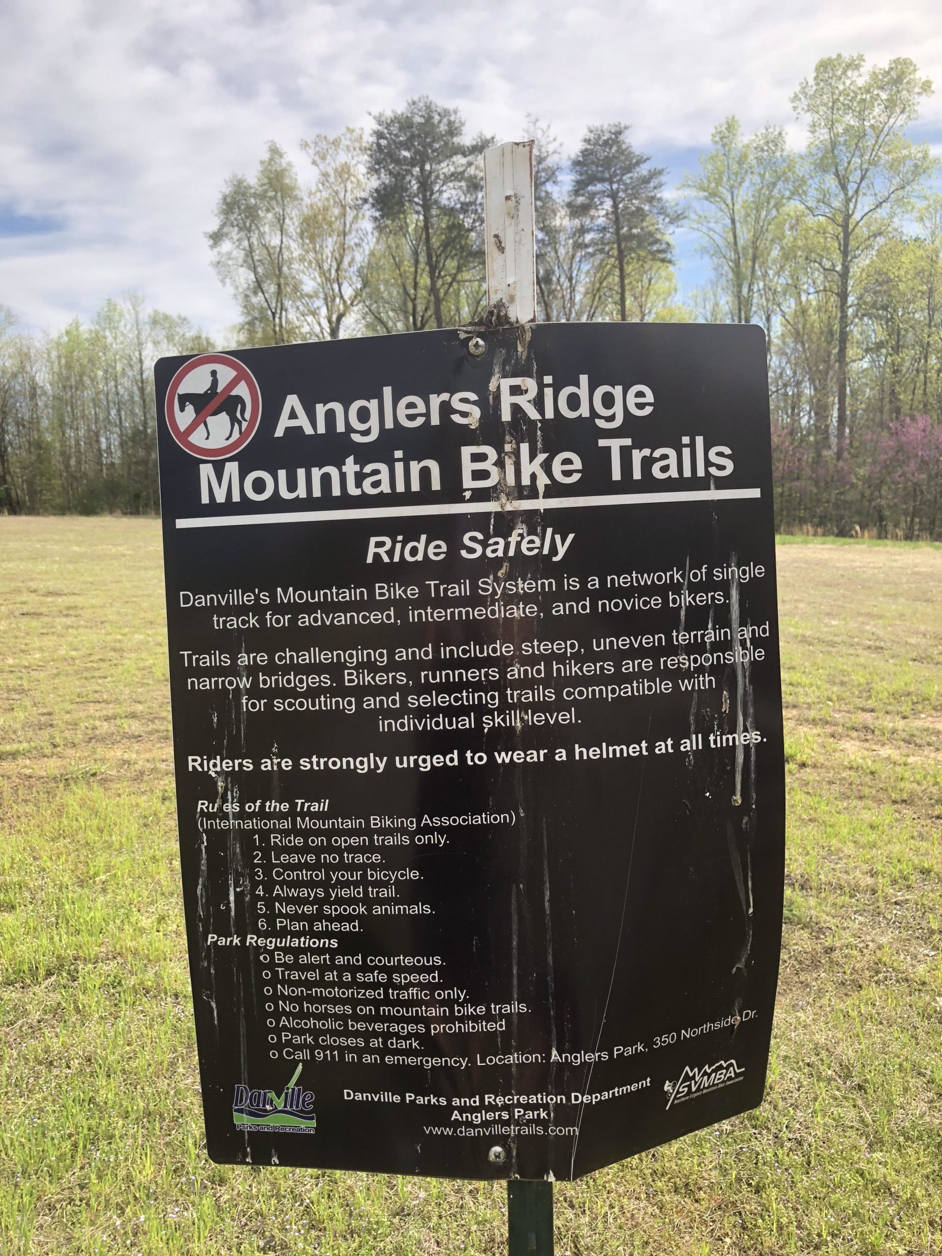Sign for Anglers Ridge Mountain Bike Trails, detailing safety information and trail regulations. The sign emphasizes the importance of helmet use, outlines rules for trail usage, and mentions that trails are suitable for various skill levels. The background features a grassy area and trees under a cloudy sky. Angler's Ridge mountain bike trail.