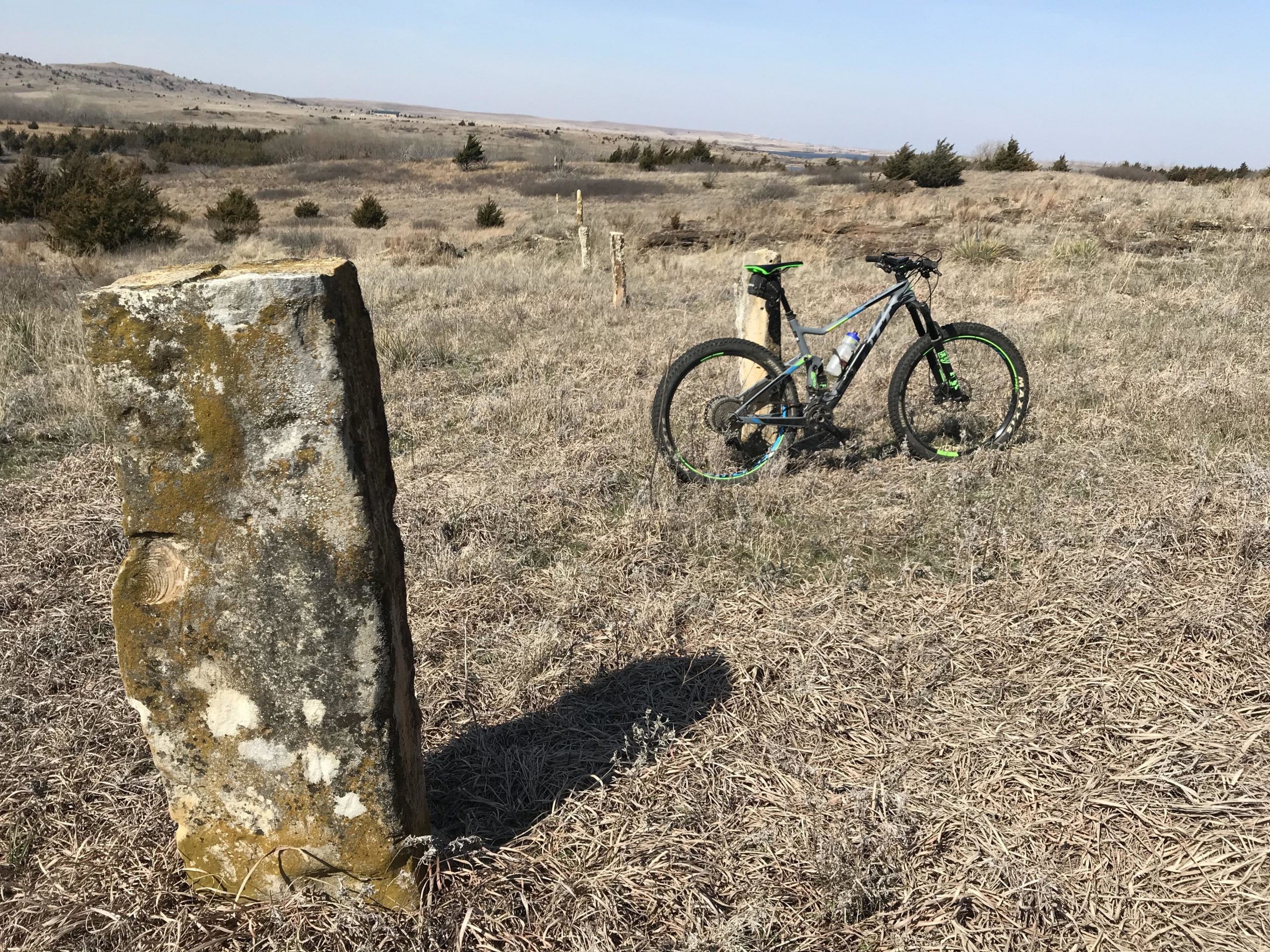 A mountain bike parked next to a weathered stone in a grassy field, with rolling hills and scattered trees in the background under a clear blue sky. Switchgrass mountain bike trail.