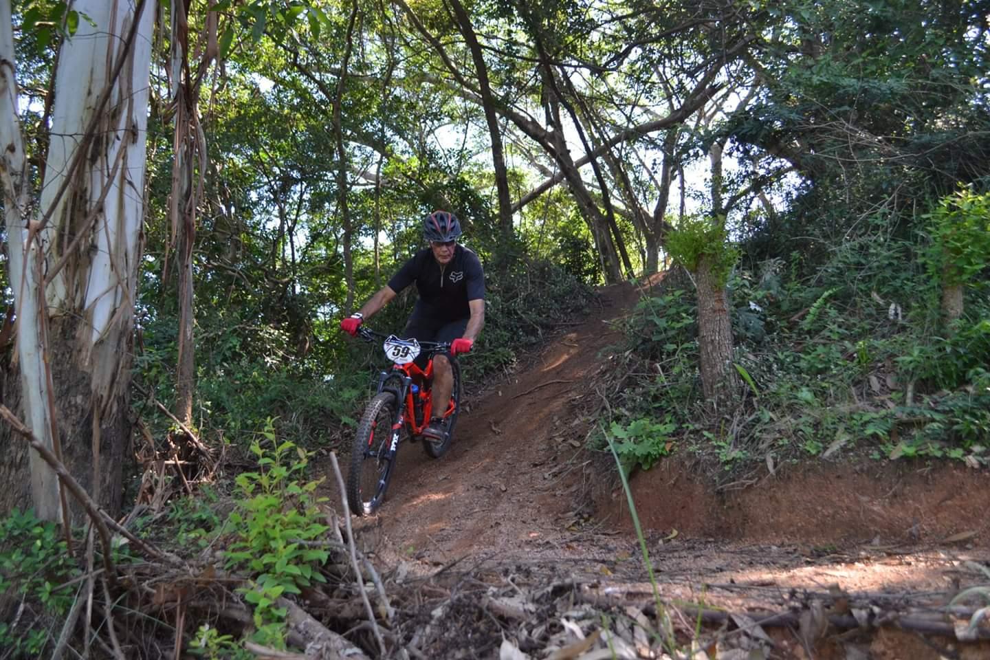 Giant Reign: A mountain biker navigating a dirt trail surrounded by lush greenery and trees, wearing a helmet and gloves, with a race number attached to the bike. The cyclist is leaning forward on a steep incline, showcasing a dynamic outdoor activity in a natural setting.