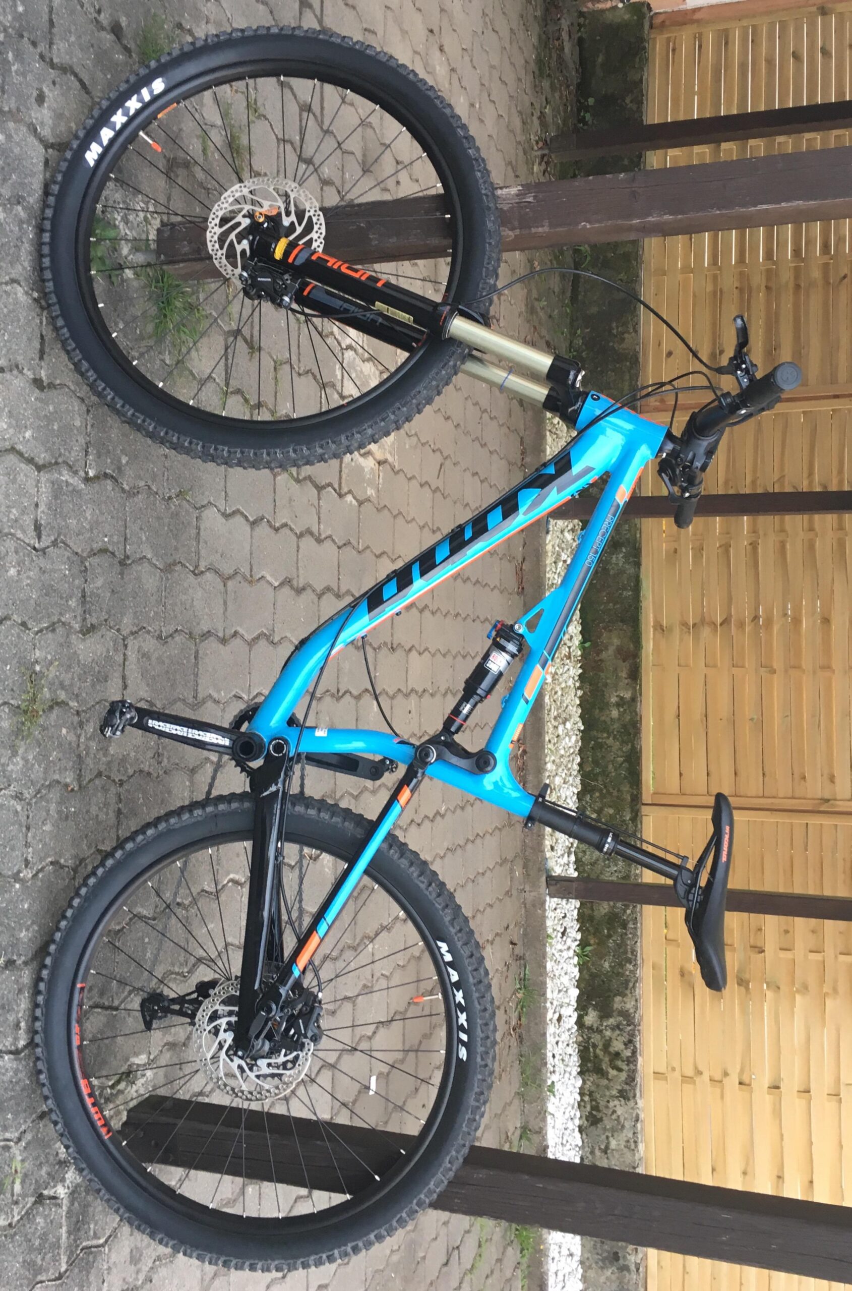 Kona Precept 130: A blue mountain bike with black tires is leaning against a wooden post. The bike features a front suspension fork and disc brakes, with visible brand markings on the tires. The background consists of a stone pavement and a wooden fence.