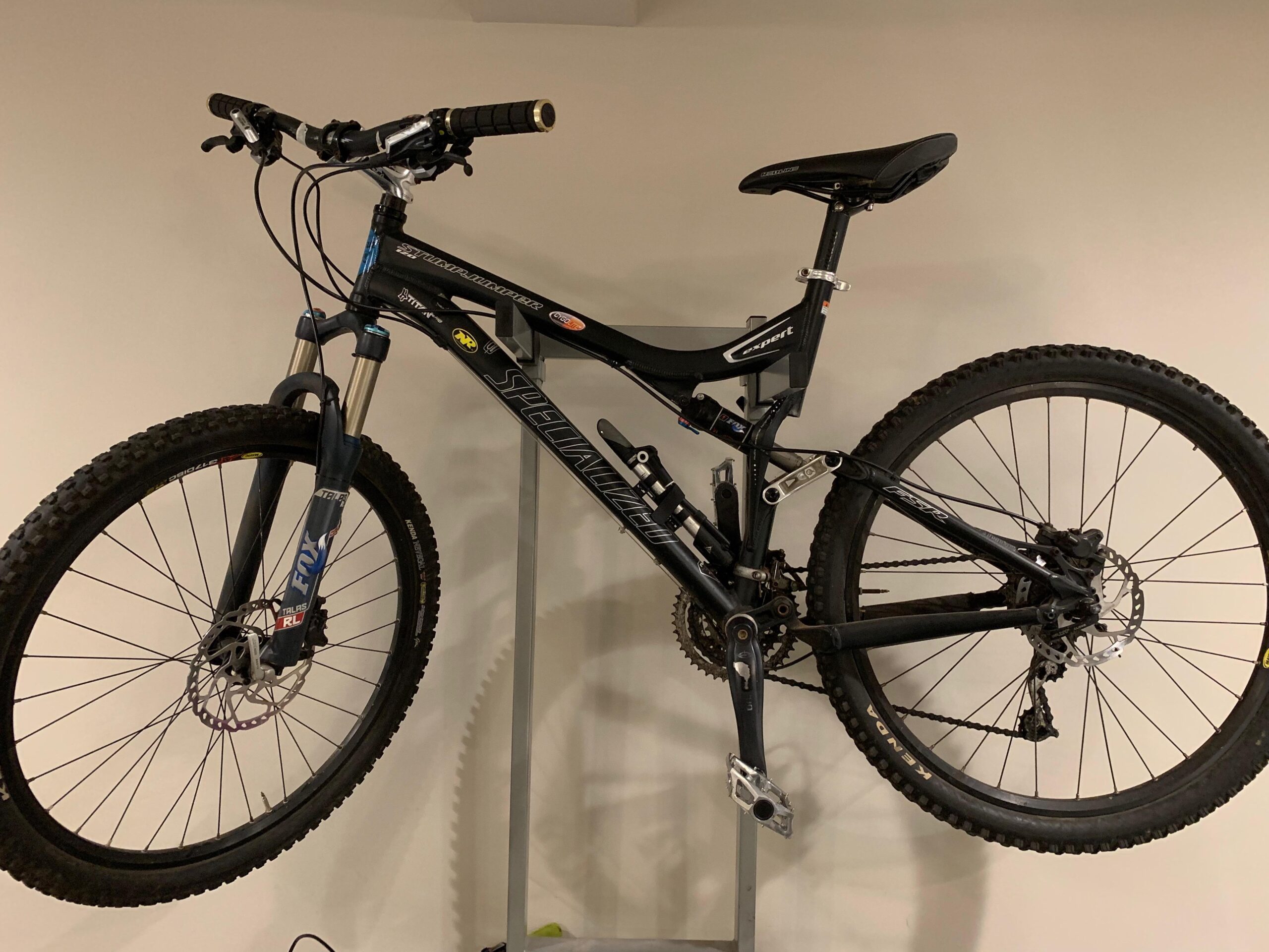 Specialized Stumpjumper FSR Expert: A black mountain bike hanging on a wall rack, featuring front and rear suspension, knobby tires, and a sleek design. The bike has black handlebars with rubber grips and a black saddle, showcasing various components such as disc brakes and a multiple-gear system. The background is a plain, light-colored wall.