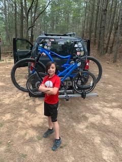 Giant Giant trance 2 29er: A young child in a red t-shirt and black shorts stands confidently in front of a vehicle parked in a wooded area. Two blue bicycles are secured on a bike rack attached to the back of the vehicle. The scene is surrounded by trees and dirt.