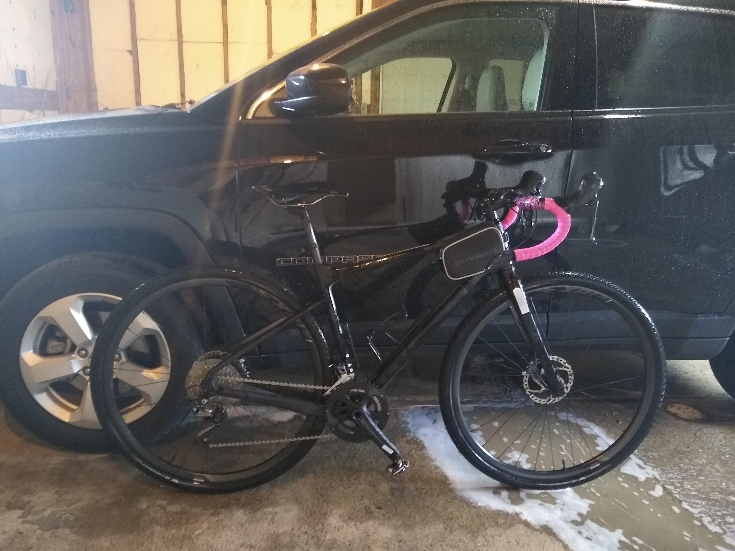 Giant Revolt 1: A black bike with pink handlebar grips parked next to a black vehicle in a garage. The bike is partially wet, and there is water on the ground. The garage walls are wooden and unfinished, indicating a work-in-progress space.