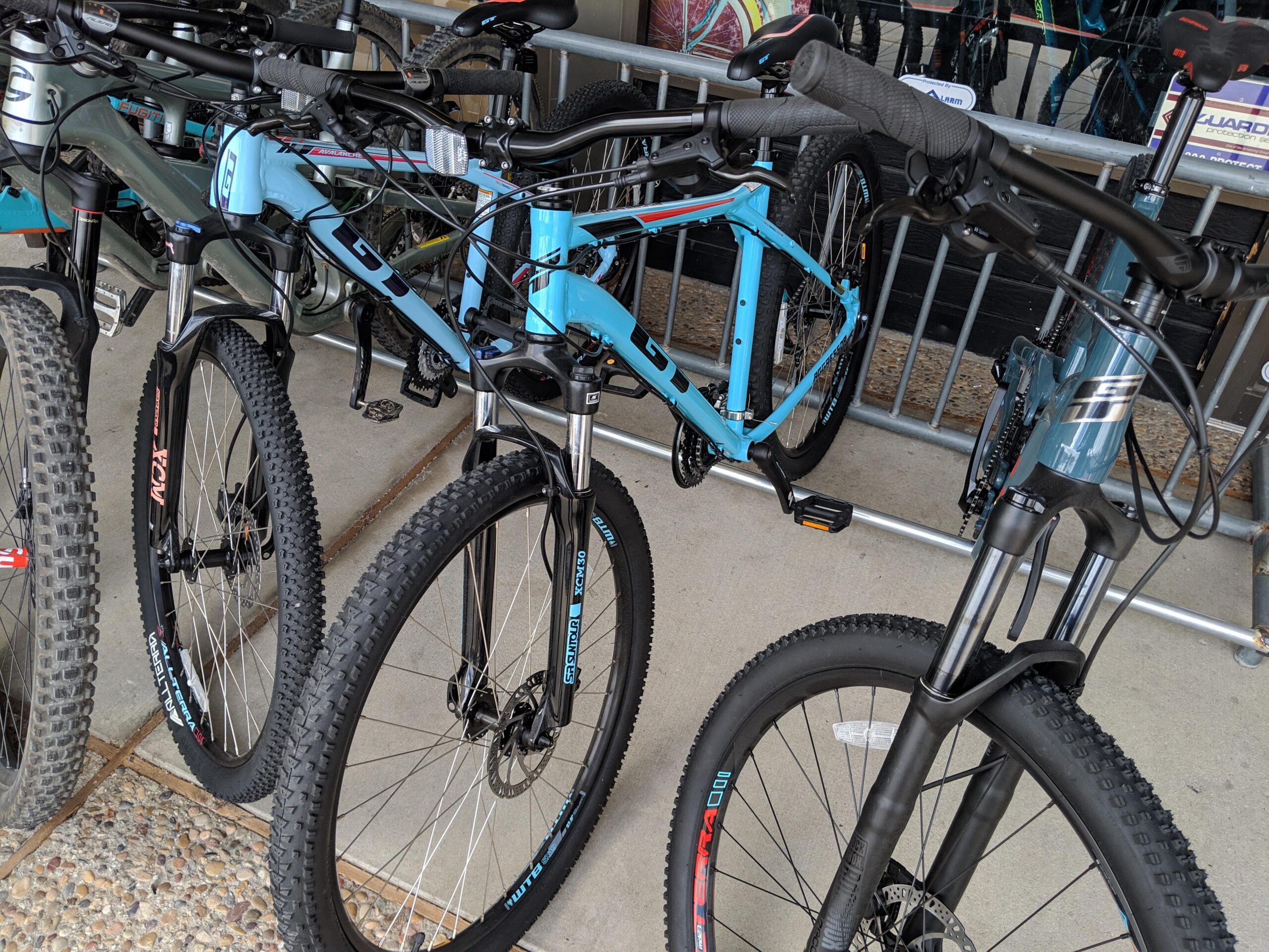 GT Aggressor: A close-up view of several bicycles parked in a bike rack. The prominent bike is a light blue mountain bike with front suspension and thick, patterned tires. Other bicycles in the background include various styles, showcasing different colors and designs. The setting appears to be a bike shop or rental area, with a hint of a reflective window displaying additional bikes.