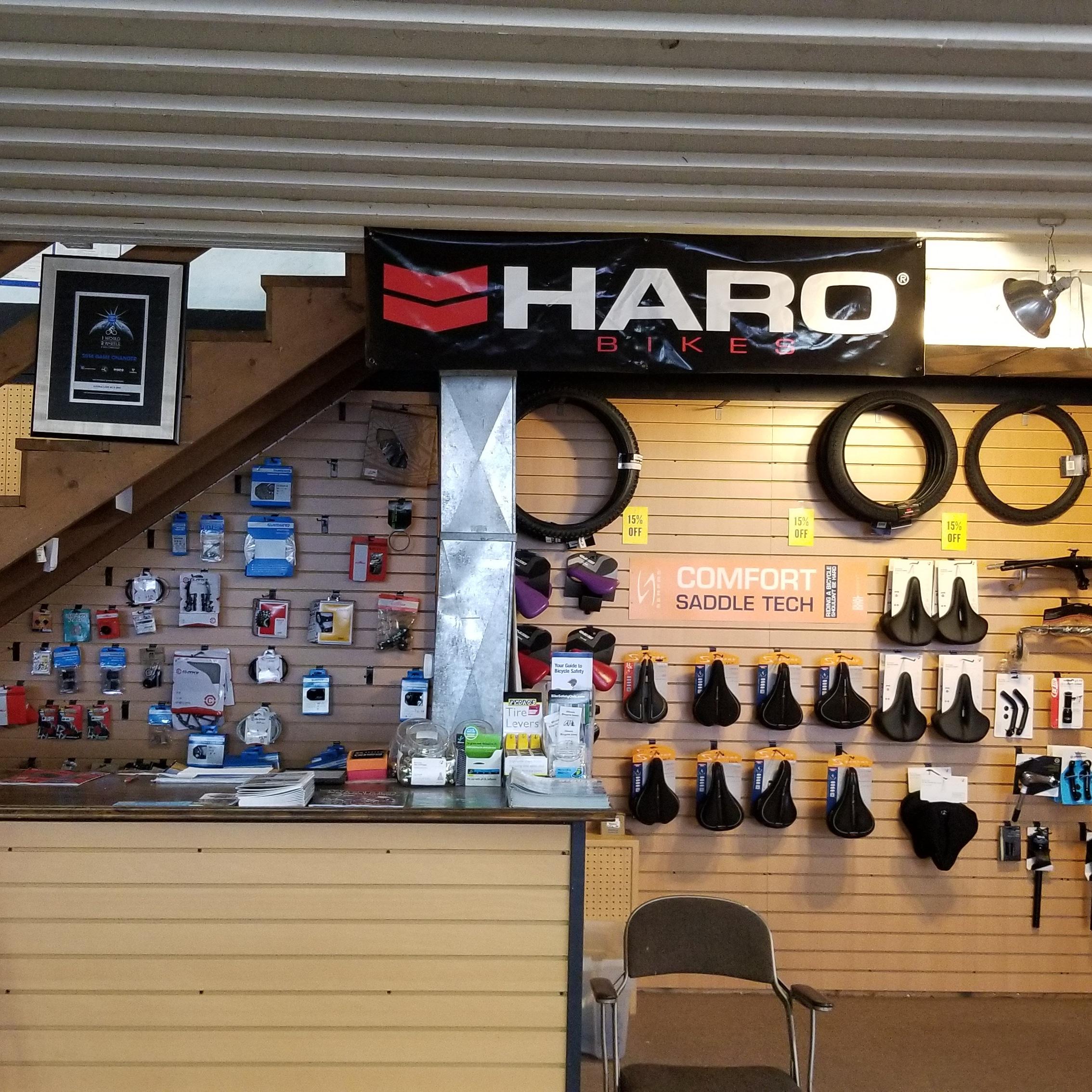 Alt text: "Interior view of a bike shop featuring a display wall with various bike accessories, seats, and tires. The shop has a counter with promotional materials, and a large banner displaying 