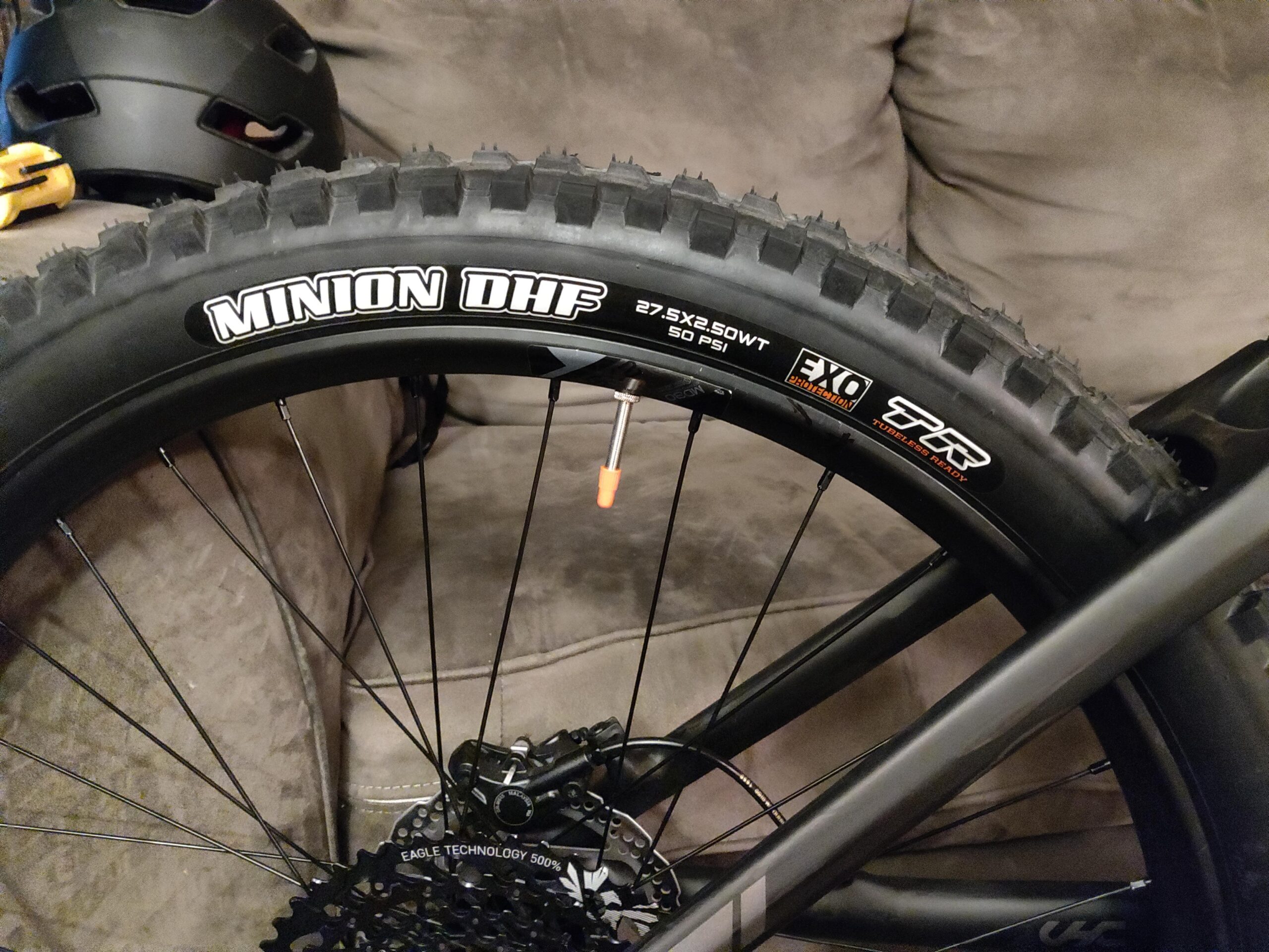 Maxxis Minion DHF: A close-up view of a mountain bike tire with the branding "MINION DHF" visible. The tire is mounted on a black wheel, and details include markings for size (27.5x2.50WT) and pressure (50 PSI). In the background, parts of a black helmet and a tool can be seen, resting on a gray couch. The image captures the bike's rugged aspect, showcasing its readiness for terrain challenges.