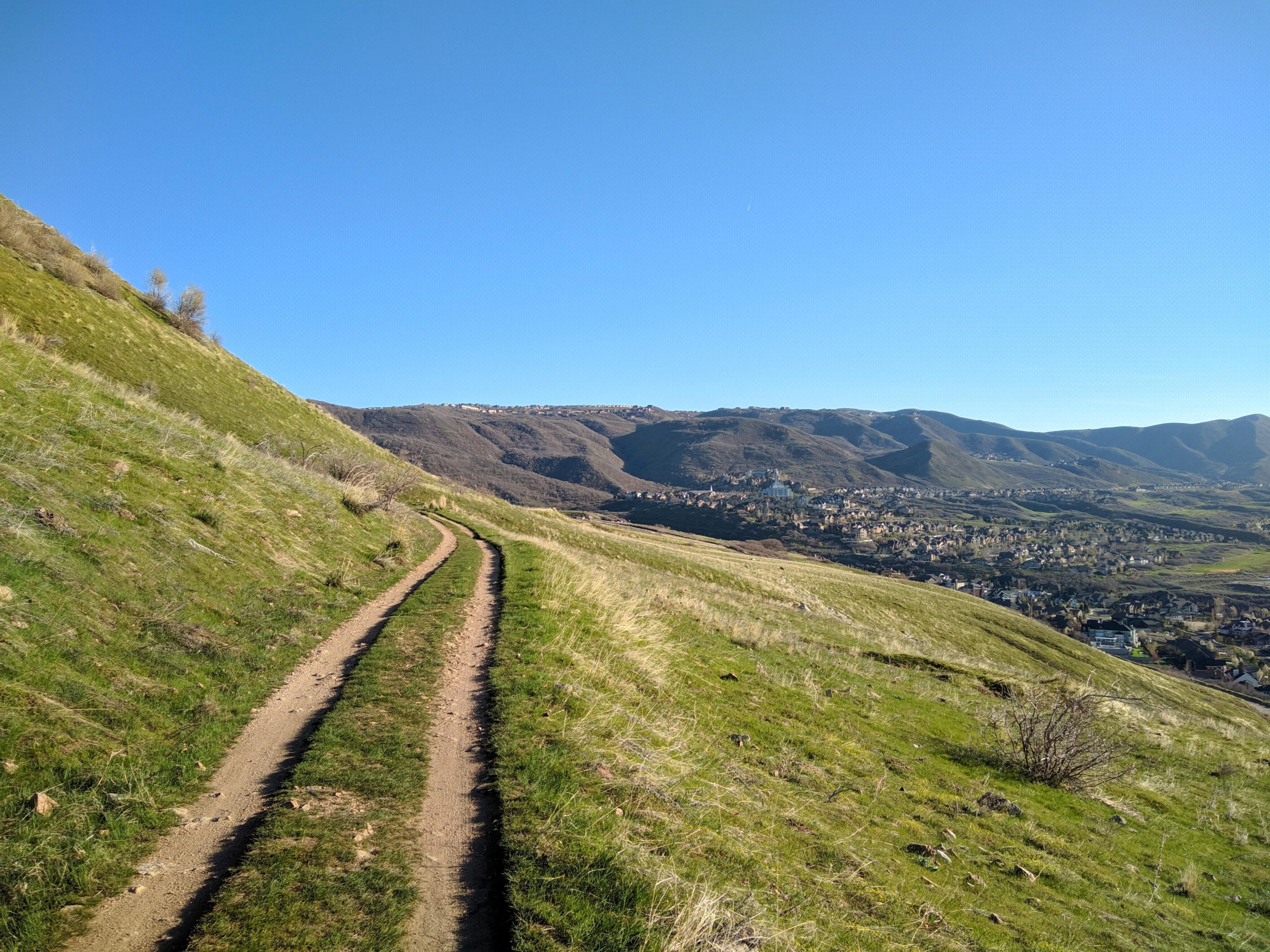 A winding dirt path traverses a grassy hillside, surrounded by lush green vegetation, leading towards a distant town nestled in a valley. The sky is clear and blue, with rolling mountains in the background. Bst To Equestrian Center Loop mountain bike trail.
