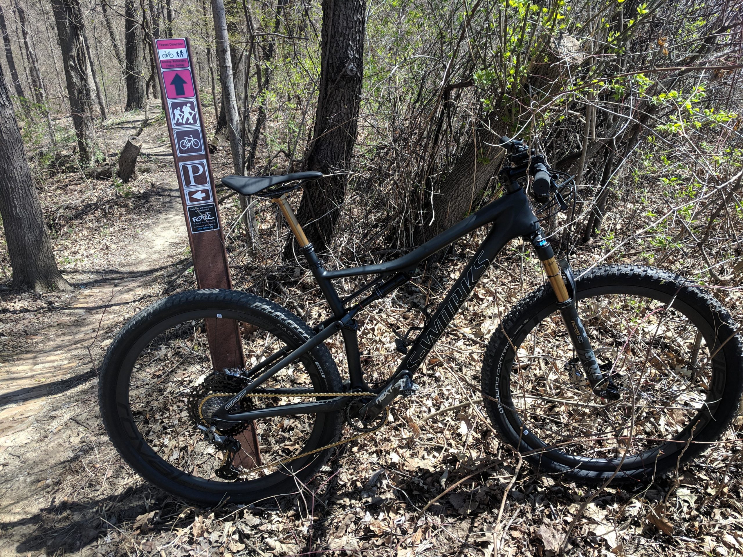 A mountain bike resting on the ground next to a trail sign in a wooded area. The sign features various icons indicating trail directions for hikers, cyclists, and parking. Surrounding the bike and sign are dry leaves and emerging greenery typical of early spring. Illiniwek Forest Preserve mountain bike trail.