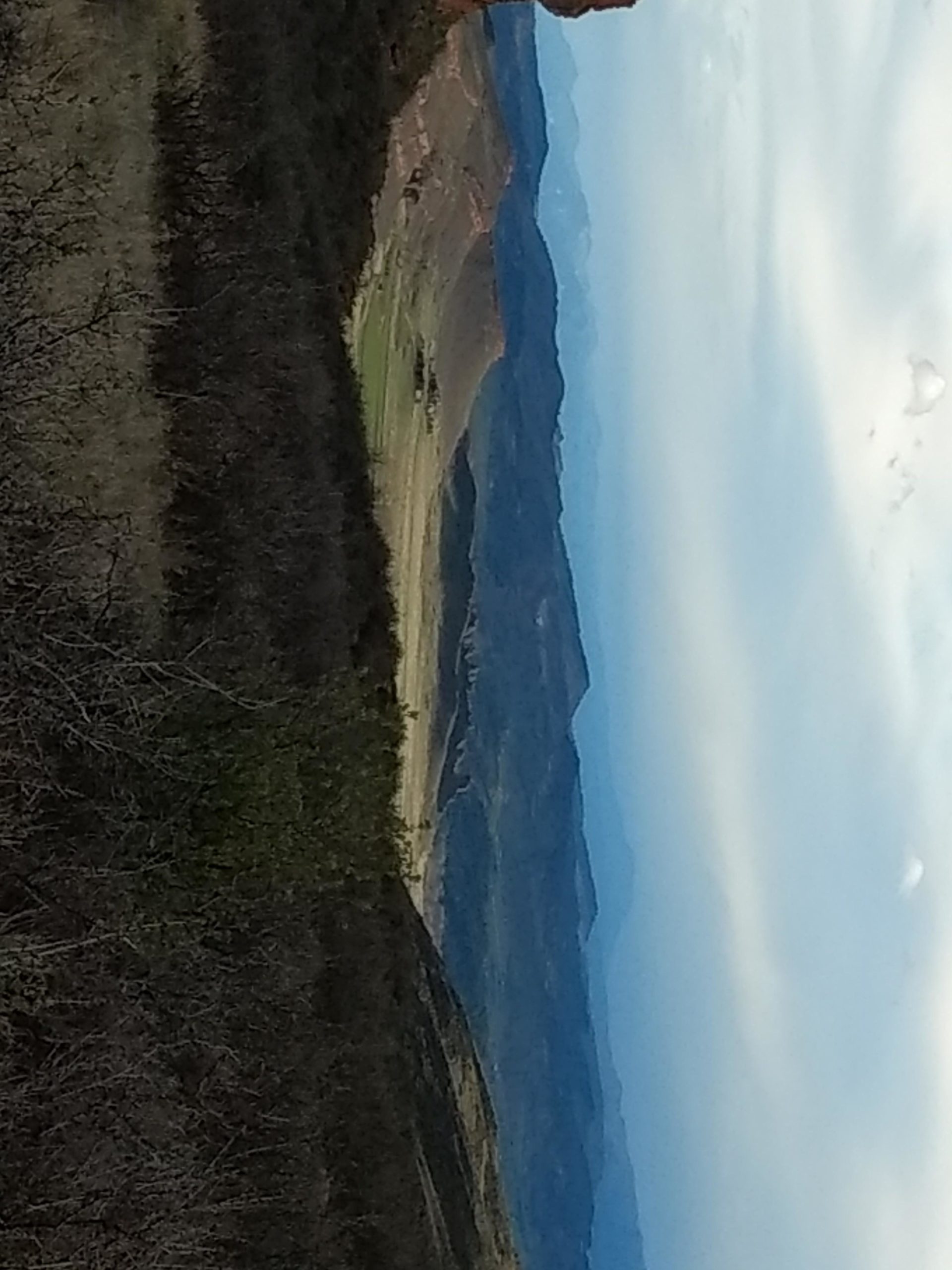 A panoramic view of rolling hills and distant mountains under a cloudy sky, with sparse vegetation in the foreground and a soft gradient of blue hues in the background. Rimrock mountain bike trail.