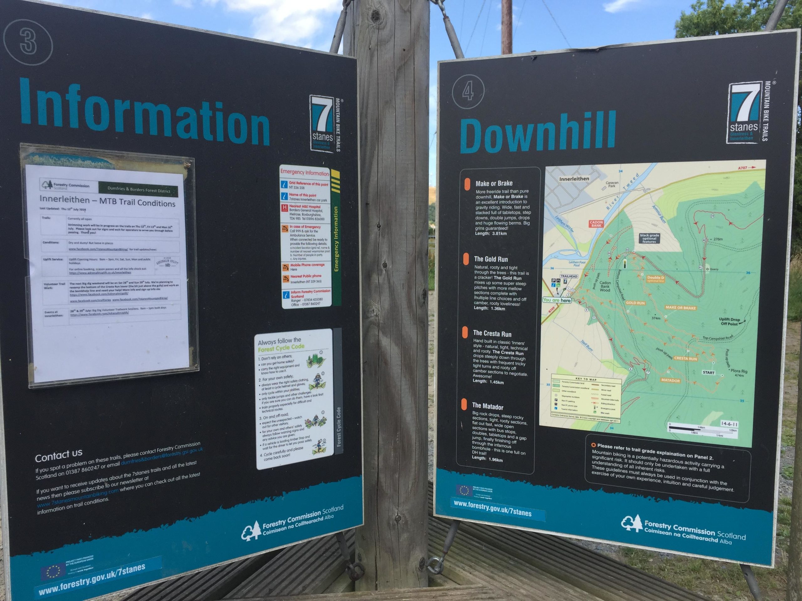 A dual-sided information sign at a mountain biking trail location in Innerleithen, Scotland. One side displays current trail conditions and emergency information, while the other side provides a detailed map of downhill biking routes, including descriptions and lengths of various trails like "Make or Brake," "The Gold Run," and "The Cresta Run." The sign features logos from forestry and cycling organizations, and includes contact information for trail issues. Innerleithen mountain bike trail.