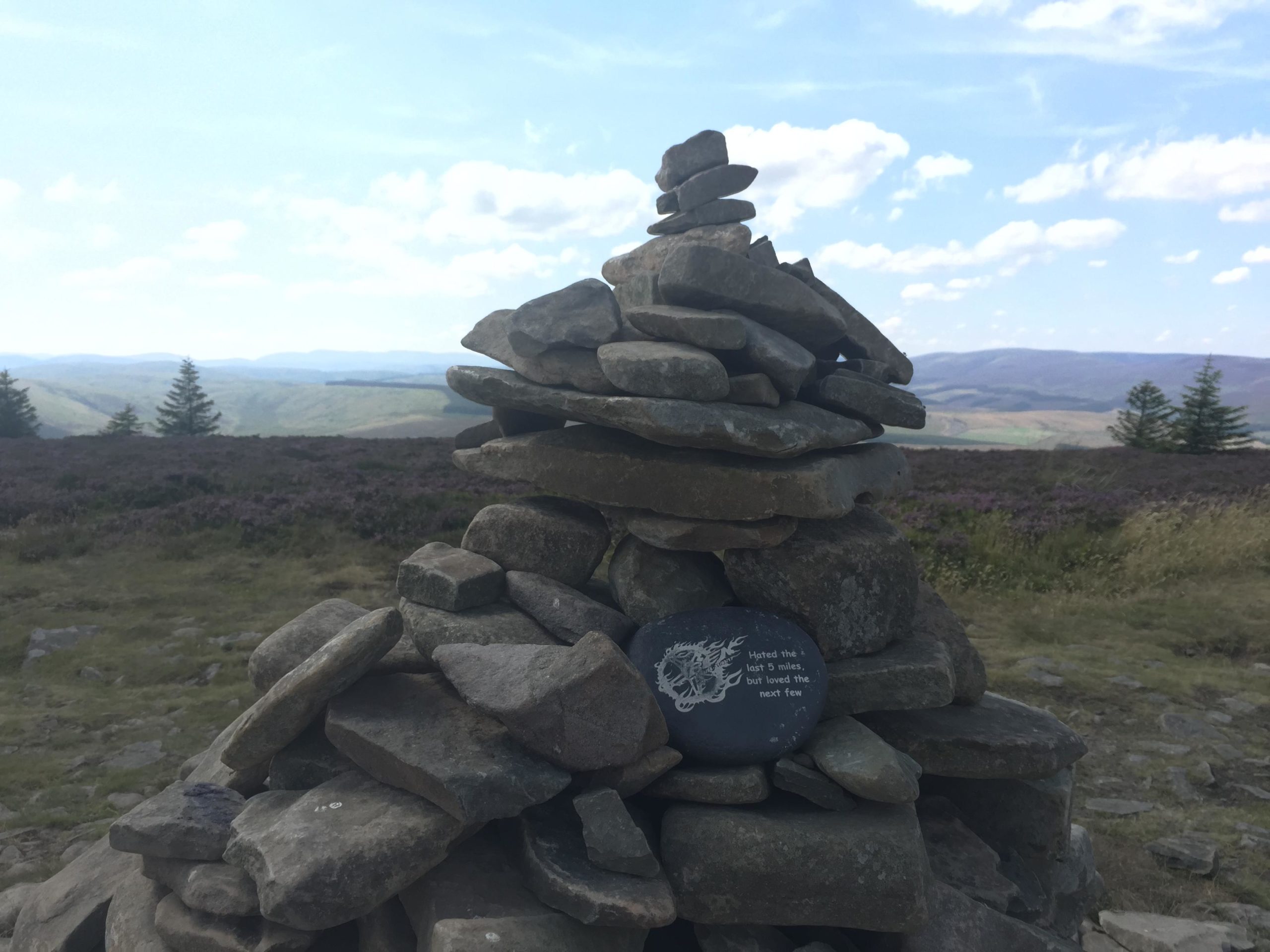 A stone cairn made of stacked rocks with a decorated black stone at its base, set against a backdrop of rolling hills and a blue sky with fluffy clouds. The area features patches of purple heather and evergreen trees in the distance, suggesting a serene outdoor landscape. Innerleithen mountain bike trail.