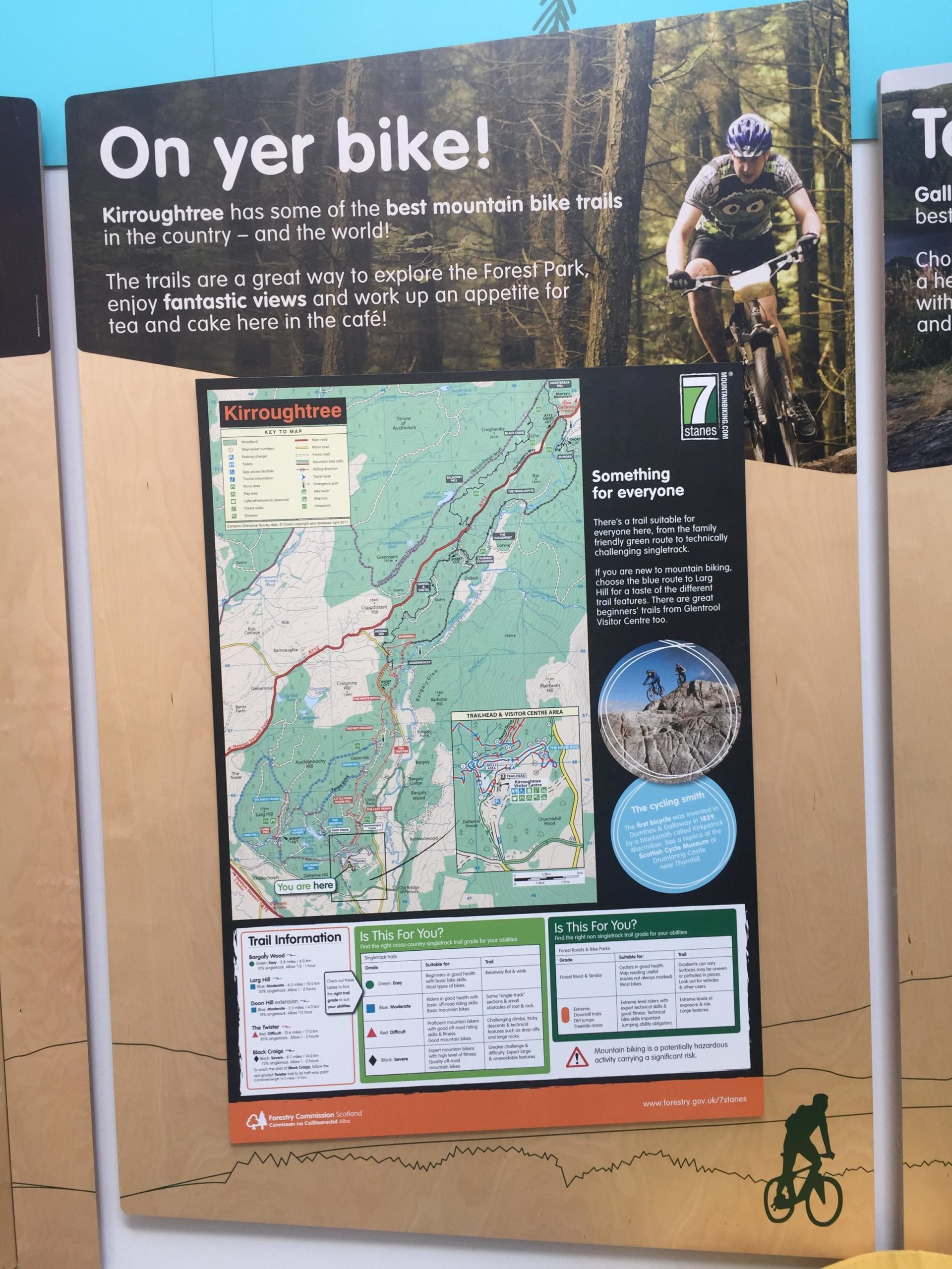 A sign promoting mountain biking in Kirroughtree, featuring a map of the bike trails in the Forest Park. The text highlights the variety of trails available for all skill levels and encourages visitors to enjoy the scenic views and local café. Images of cyclists biking are included, and there's important trail information and a warning about the nature of mountain biking. Kirroughtree mountain bike trail.