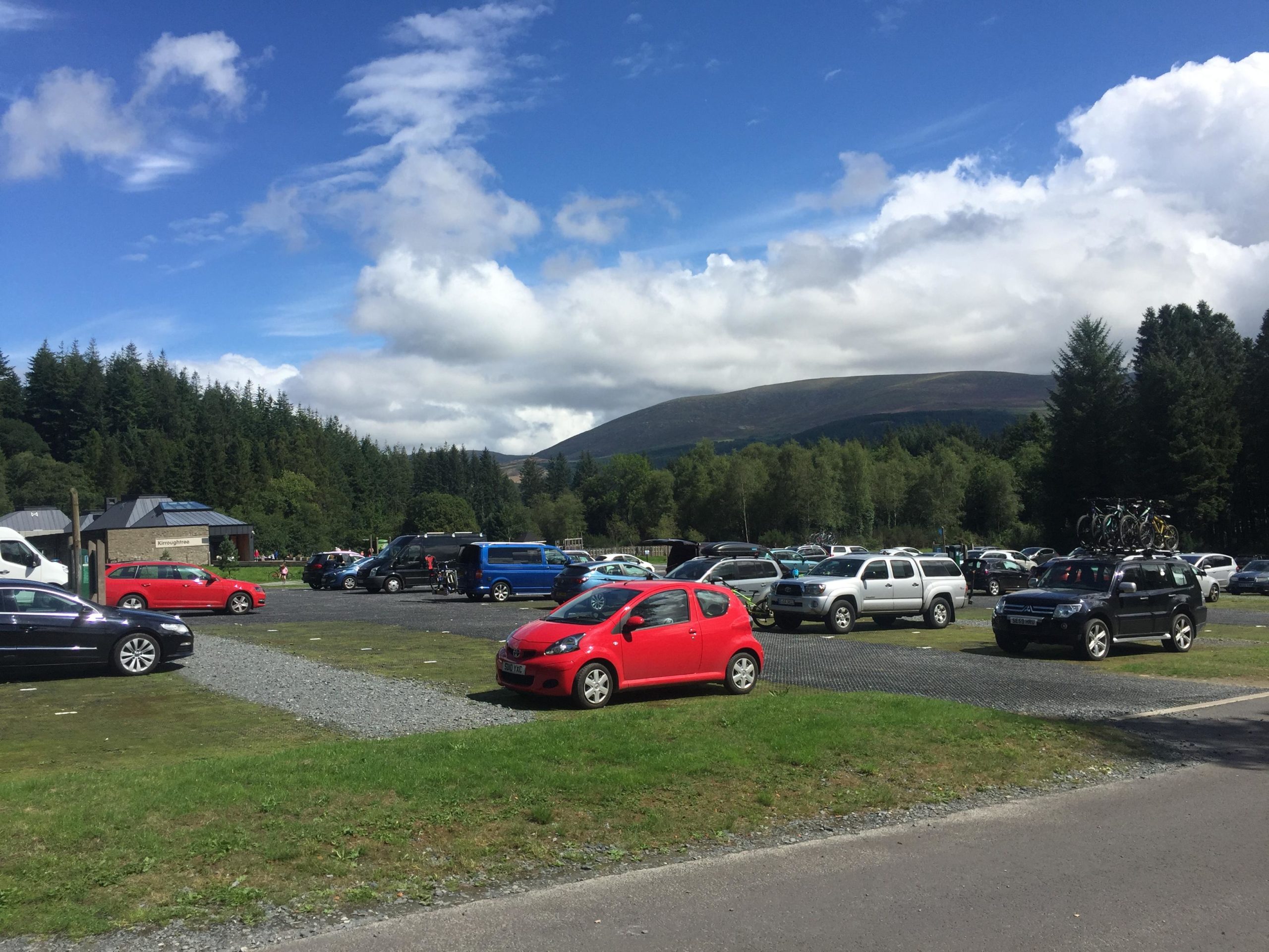A large parking area filled with various vehicles, including red cars and SUVs, set against a backdrop of green trees and distant mountains under a partly cloudy blue sky. A building is visible in the background, surrounded by nature. Kirroughtree mountain bike trail.