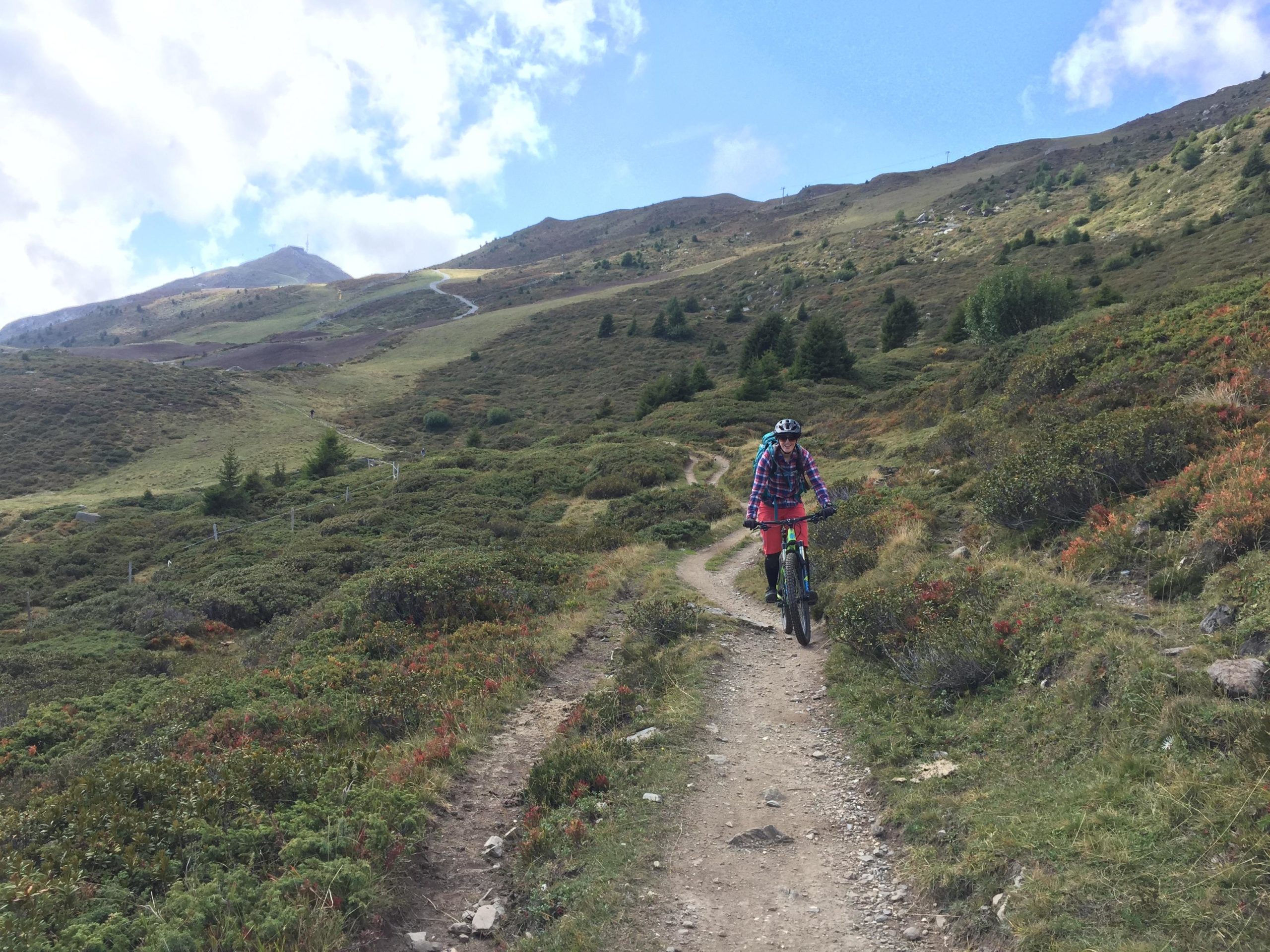A mountain biker riding along a dirt trail in a scenic landscape with rolling hills and greenery. The sky is partly cloudy, and the terrain features patches of grass and shrubs, with a mountain visible in the background. Churwalden Zone mountain bike trail.