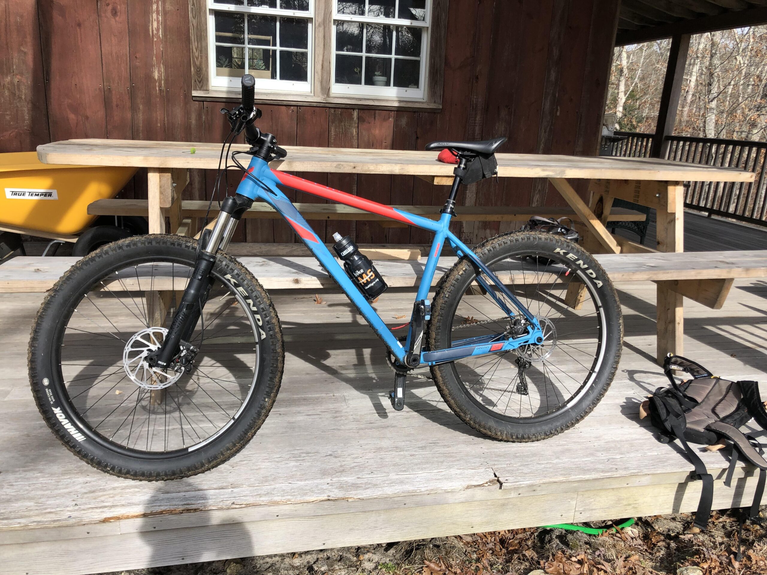 Devinci Devinci Kobain RS: A blue and red mountain bike is parked on a wooden deck beside a picnic table, with a yellow wheelbarrow in the background. A water bottle is attached to the frame, and a small backpack is resting on the deck nearby. Soft sunlight illuminates the scene.