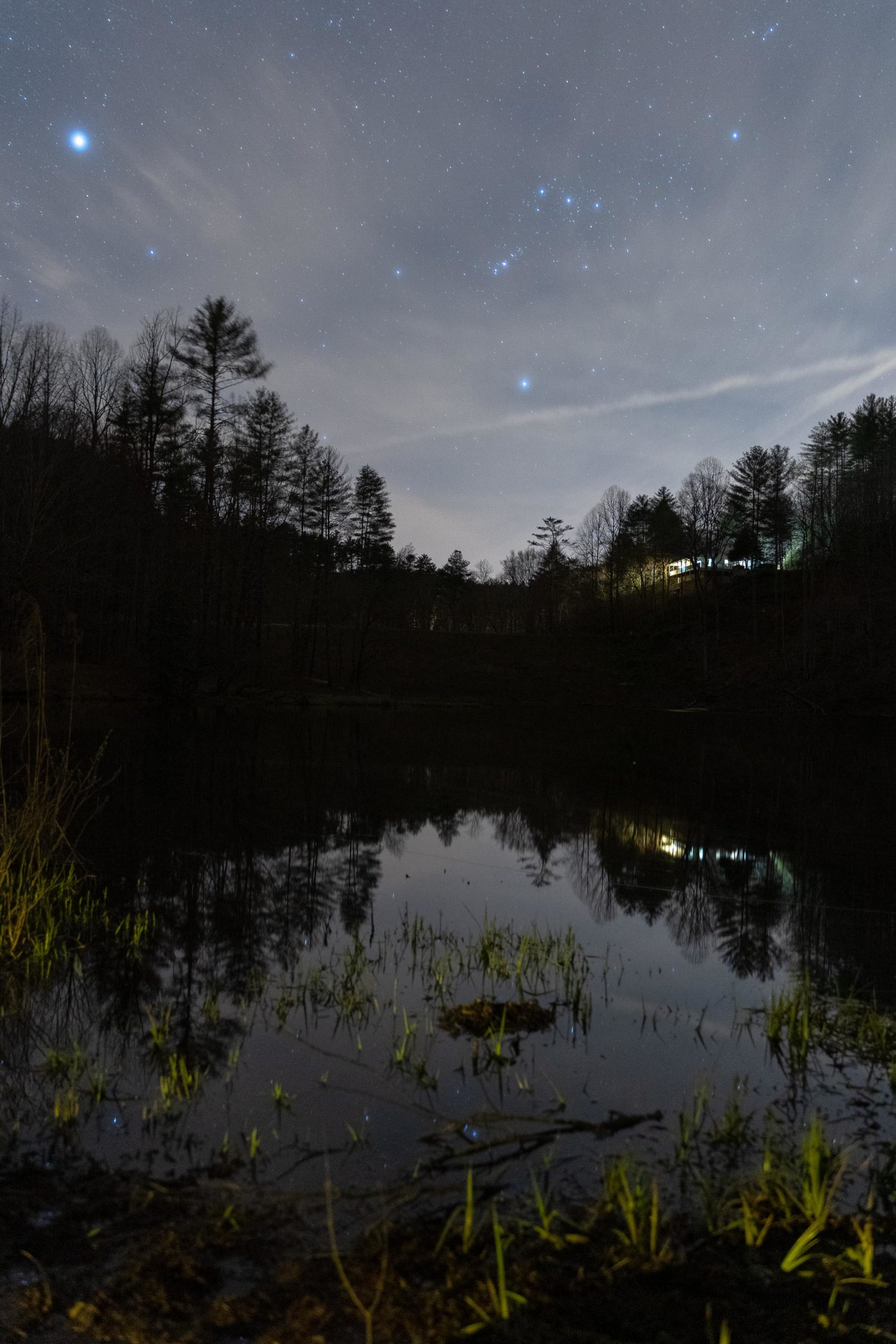 A serene nighttime landscape featuring a calm lake reflecting a starry sky. Silhouetted trees line the background, with some faint clouds. A small, illuminated structure can be seen on a hill, adding a warm glow to the scene. Grass and reeds emerge at the water's edge, creating a tranquil atmosphere. Pinhoti Trail: Mountaintown Creek Segment mountain bike trail.