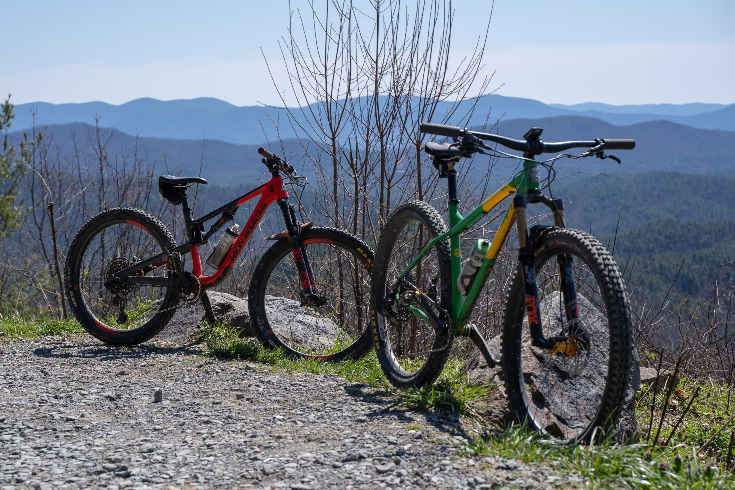 Two mountain bikes, one red and one green, are positioned on a gravel path overlooking a mountainous landscape. The bikes are parked beside a rocky outcrop, with sparse trees in the background and a clear blue sky above. The scene captures the essence of outdoor adventure and cycling. Pinhoti Trail: P1 / Bear Creek Pinhoti mountain bike trail.