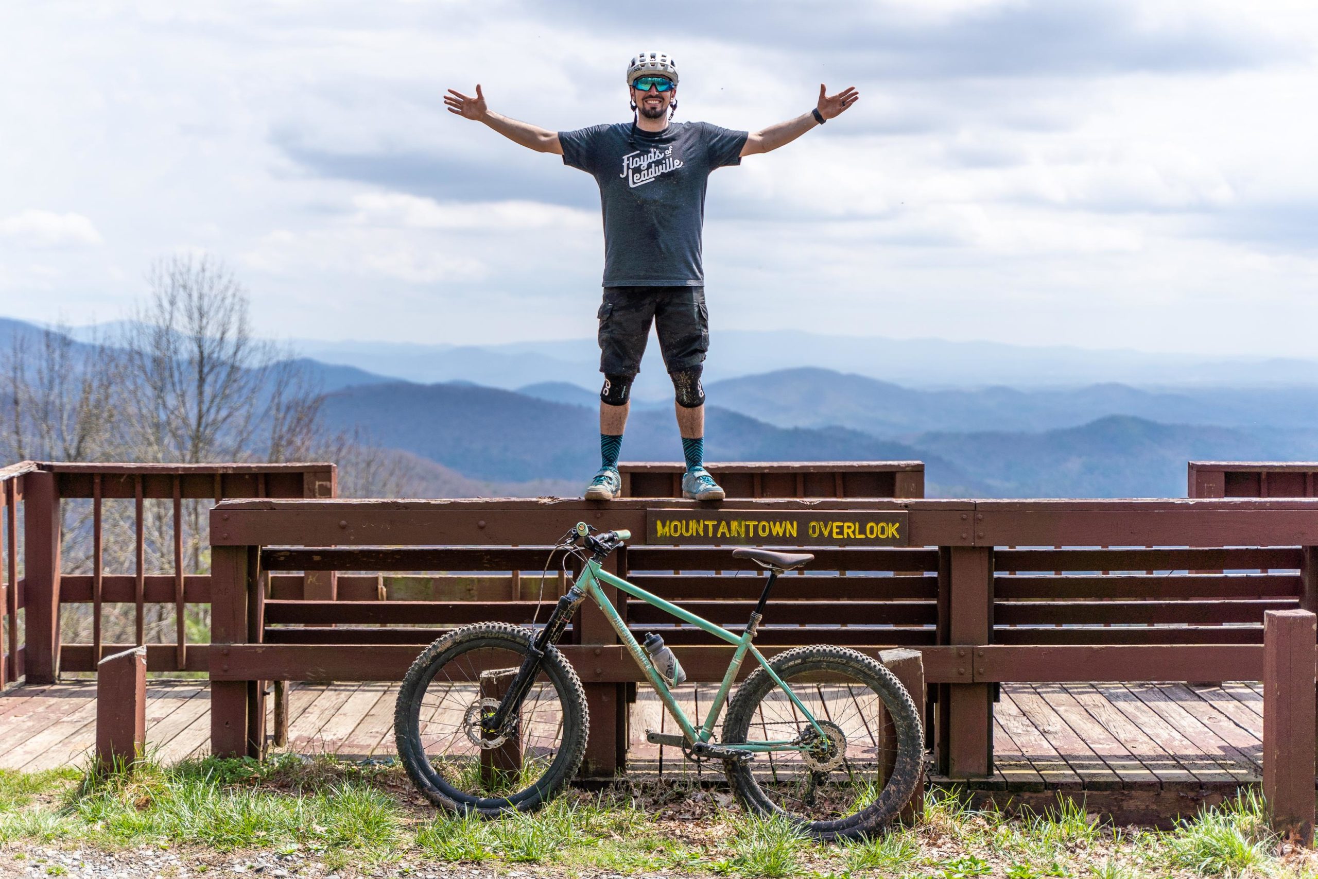 A person standing with arms outstretched on a wooden overlook with a yellow sign reading "Mountaintown Overlook." In the foreground, a mountain bike leans against the railing. The background features rolling mountains under a cloudy sky. The scene conveys a sense of adventure and triumph in nature. Pinhoti Trail: Mountaintown Creek Segment mountain bike trail.