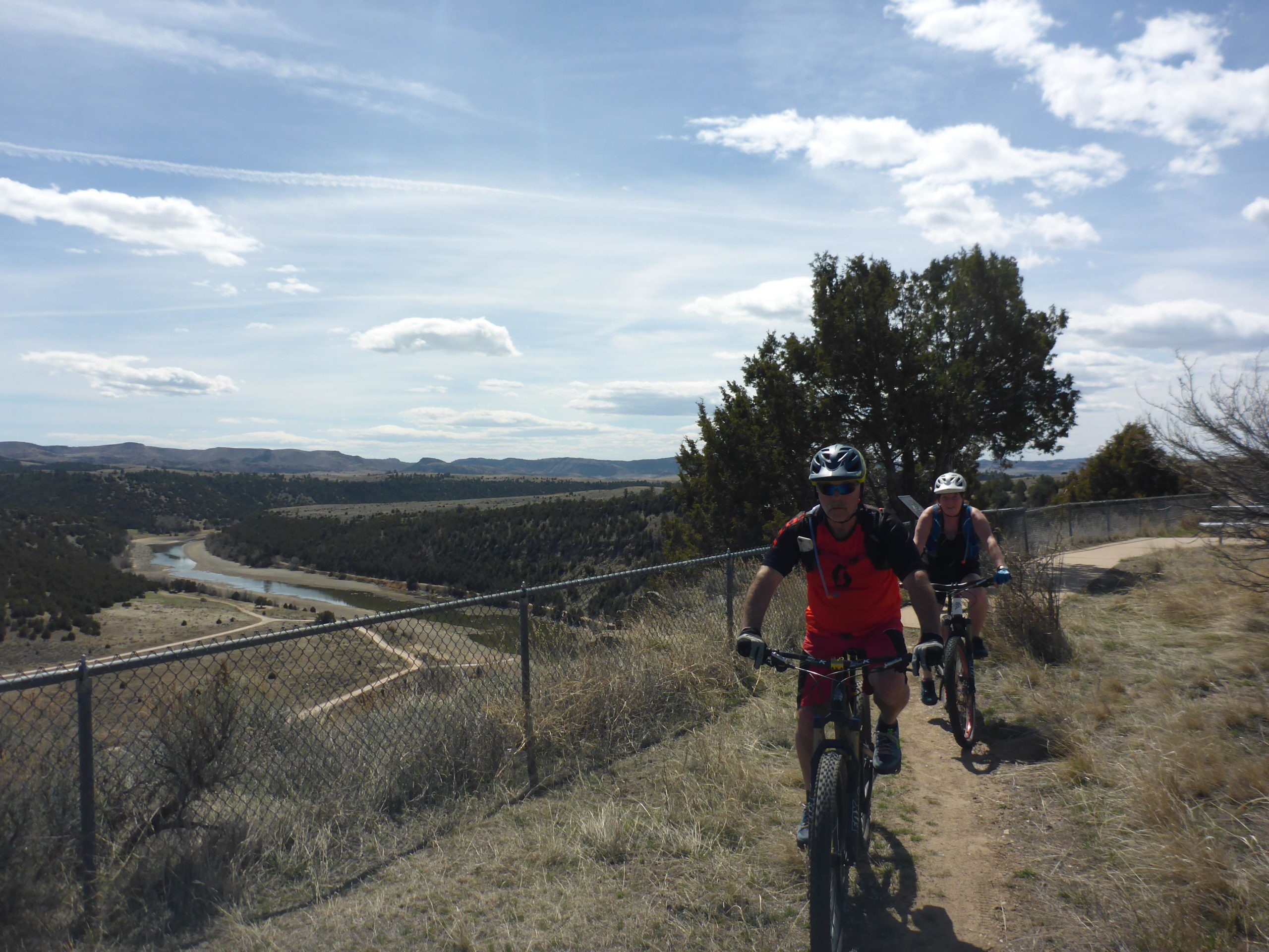 Two mountain bikers navigate a dirt trail along a scenic overlook, with a winding river and grassy hills in the background. The sky is partly cloudy, and there are patches of greenery and sparse trees along the trail. The trail is bordered by a chain-link fence. Glendo State Park mountain bike trail.