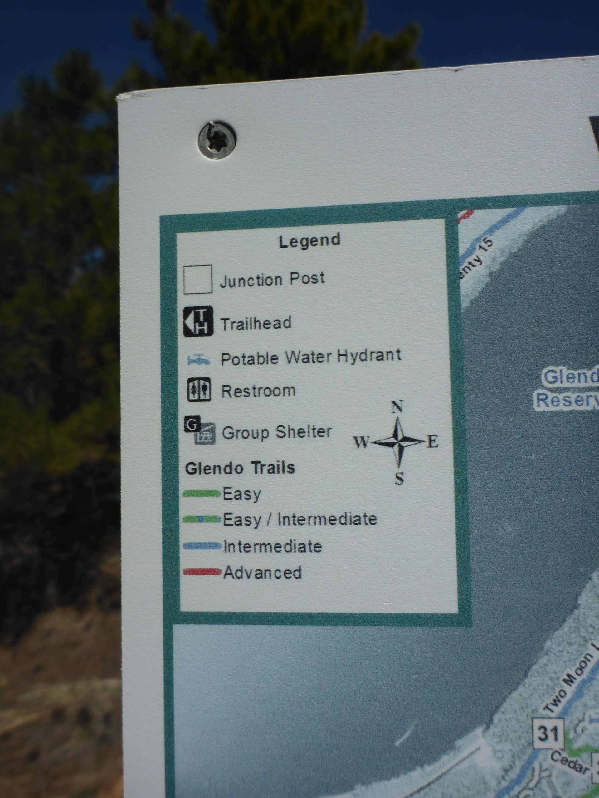 A close-up of a trail map sign featuring a legend that indicates various symbols for Junction Post, Trailhead, Potable Water Hydrant, Restroom, and Group Shelter. The map also categorizes Glendo Trails by difficulty levels: Easy, Easy/Intermediate, Intermediate, and Advanced, along with a compass rose showing north, south, east, and west directions. Glendo State Park mountain bike trail.