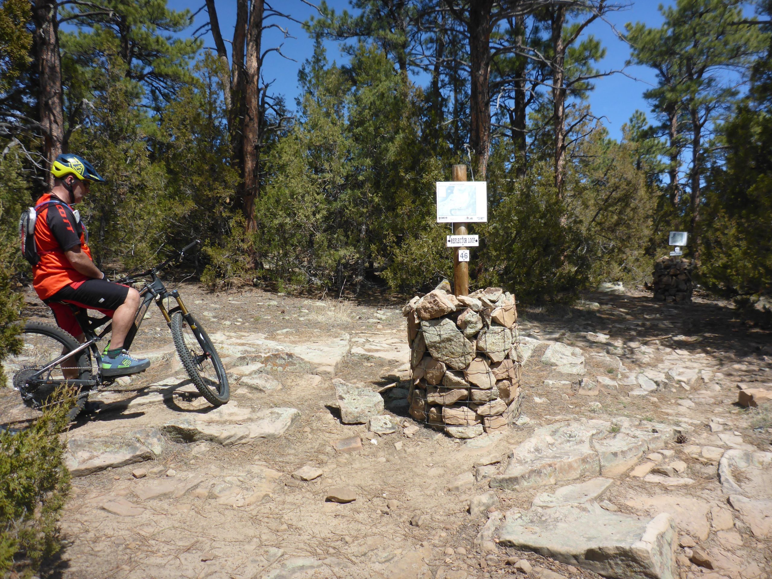 A mountain biker in an orange jersey and helmet pauses beside a rocky trail junction marked by two signs on stone pillars, surrounded by pine trees and a clear blue sky. Glendo State Park mountain bike trail.