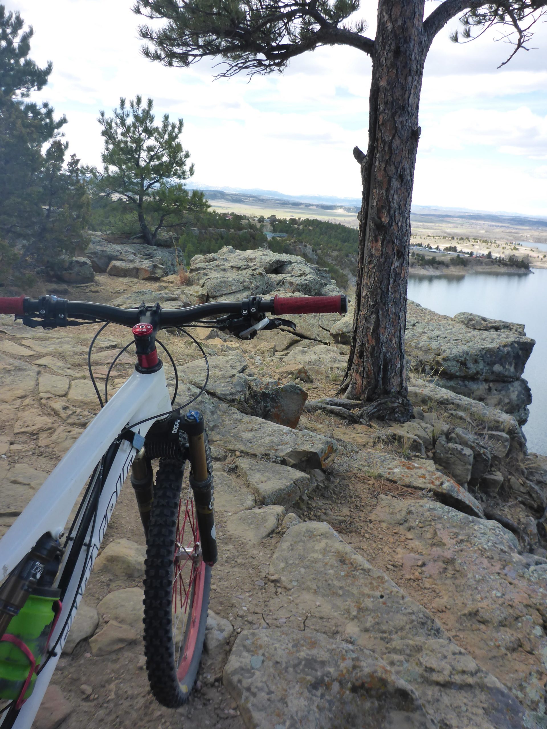 Mountain bike parked near the edge of a rocky cliff, with a view of a lake and distant mountains under a cloudy sky. Pine trees are visible in the background. Glendo State Park mountain bike trail.
