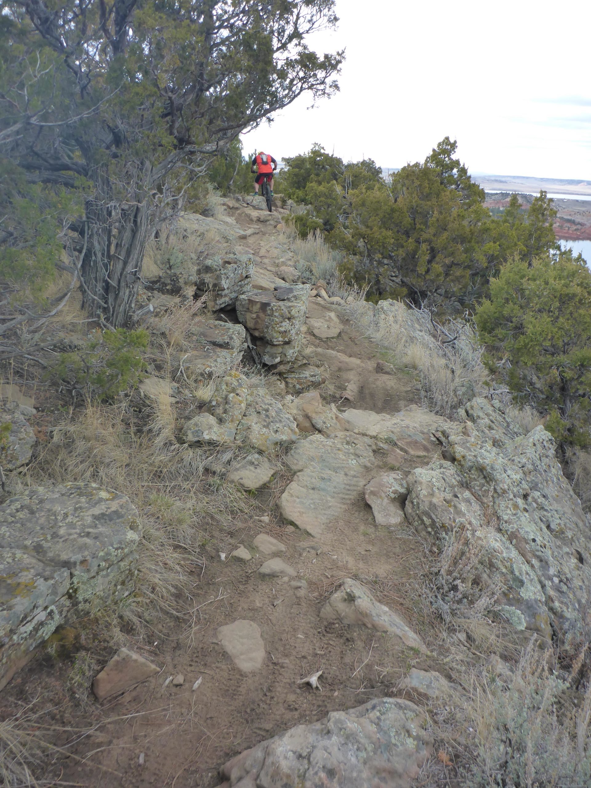 A rocky hiking trail winding through a rugged landscape, framed by shrubs and trees. A person in a red backpack is walking along the path, with a scenic view of a body of water and distant terrain visible in the background. The trail is uneven, with scattered rocks and dirt, indicating a natural setting ideal for outdoor activities. Glendo State Park mountain bike trail.