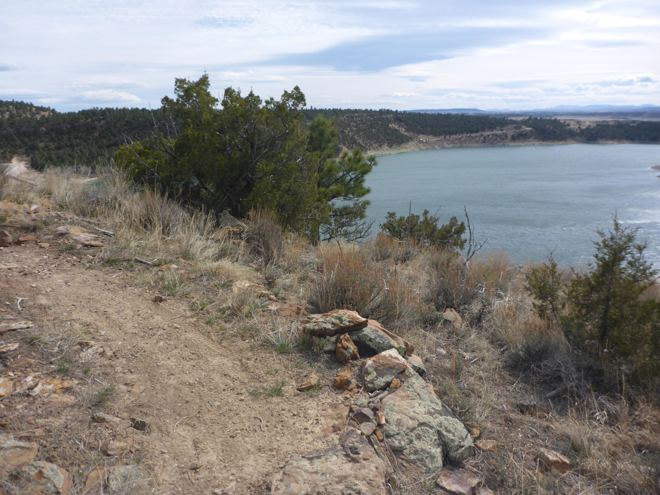 A rocky hillside overlooking a body of water, surrounded by trees and shrubs under a cloudy sky. A dirt path leads toward the edge of the slope, with patches of grass and small stones visible. Glendo State Park mountain bike trail.