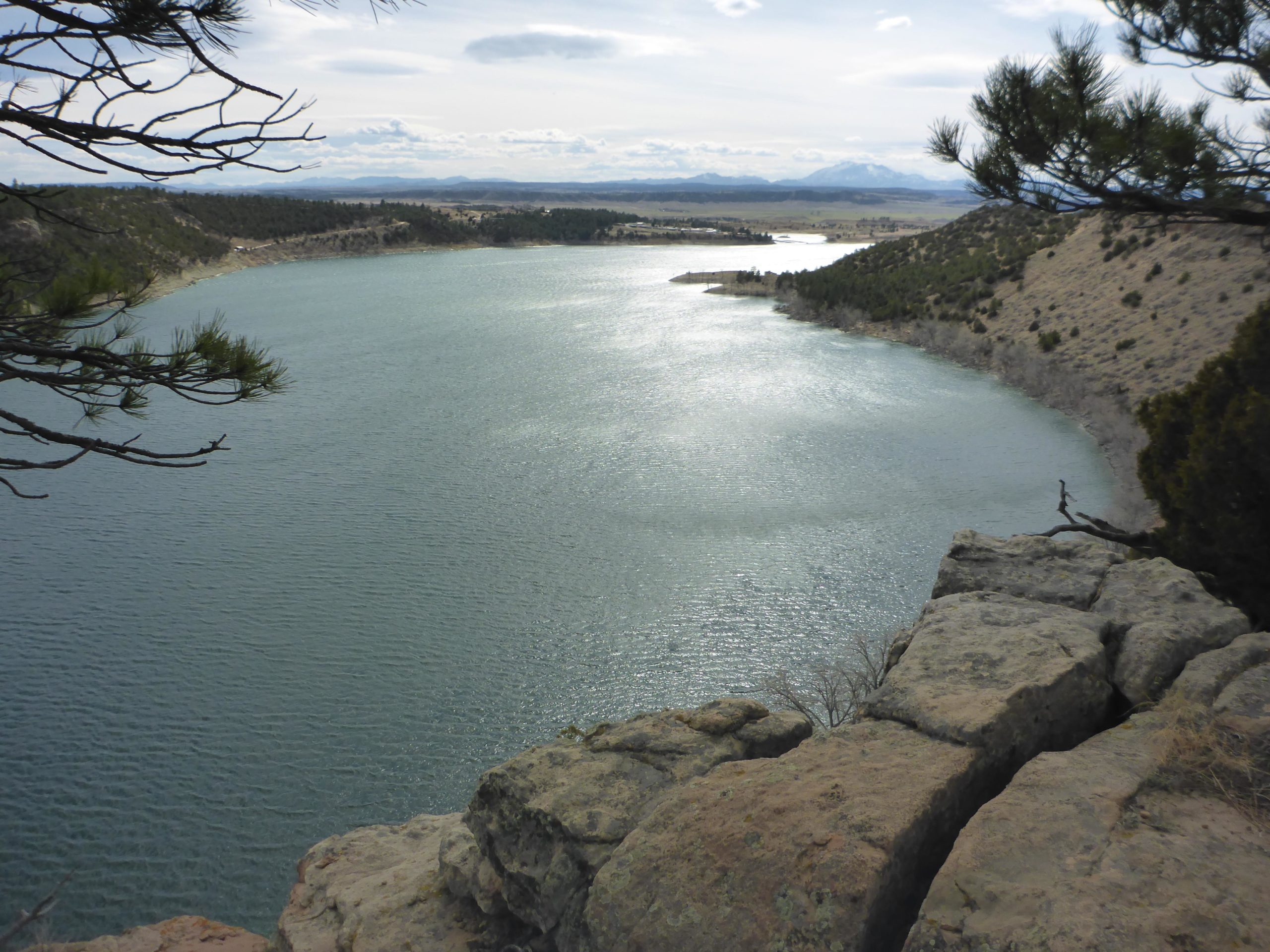 A scenic view of a calm lake surrounded by rolling hills and distant mountains, captured from a rocky cliff. The surface of the water reflects the sky, creating a serene atmosphere. Sparse trees frame the image, enhancing the natural beauty of the landscape. Glendo State Park mountain bike trail.
