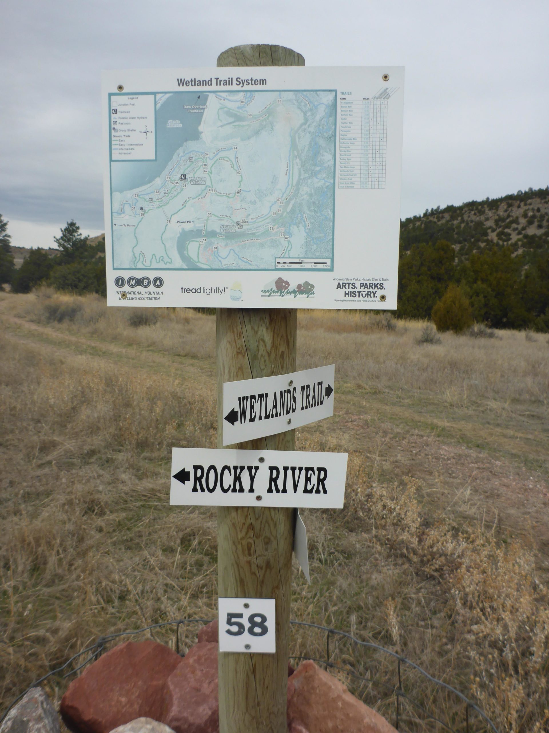 Trail signpost featuring a map of the Wetland Trail System, with directional arrows indicating "Wetland Trail" and "Rocky River," and a marker labeled "58," set against a backdrop of grassy terrain and trees under a cloudy sky. Glendo State Park mountain bike trail.