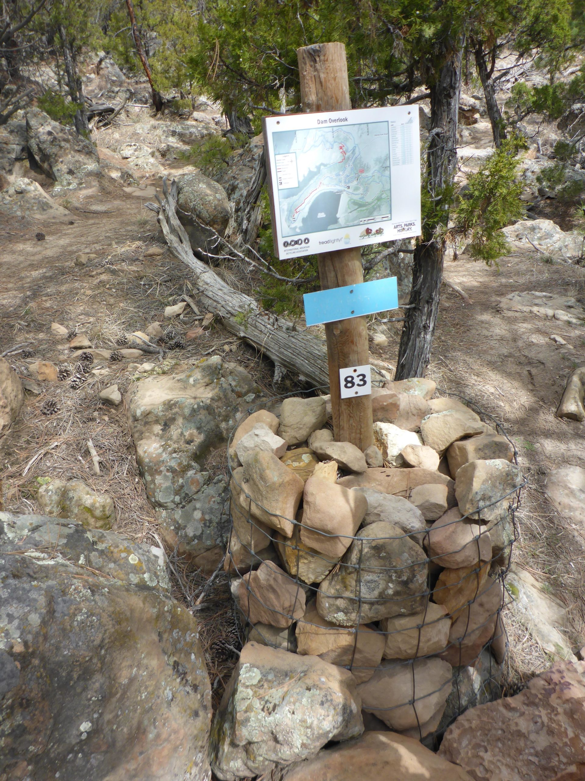 Trail signpost indicating the "Dam Overlook" along a rocky hiking path. The sign features a map with marked trails, set on a wooden post surrounded by a stone structure. The area is lined with trees and rocky terrain, suggesting a natural outdoor setting. Glendo State Park mountain bike trail.