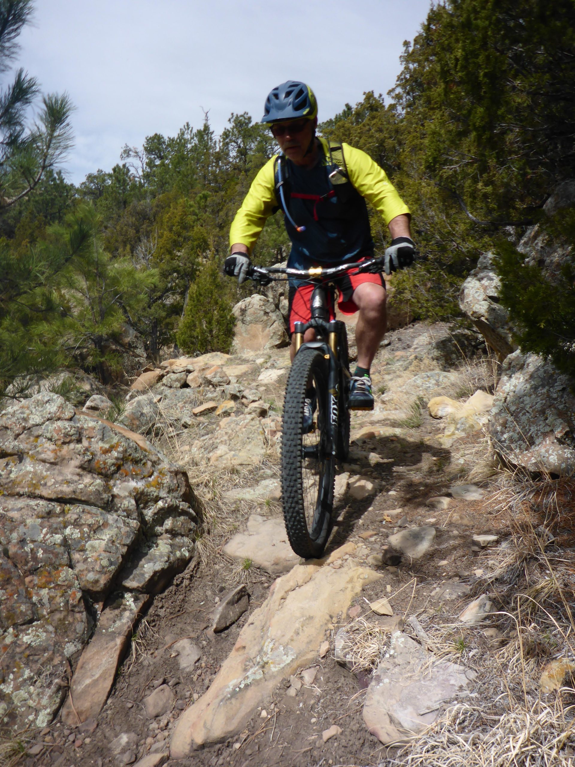 A person wearing a helmet and bright clothing rides a mountain bike along a rocky trail surrounded by trees and shrubs. The terrain features large boulders and loose stones, indicating a challenging path for mountain biking. Glendo State Park mountain bike trail.