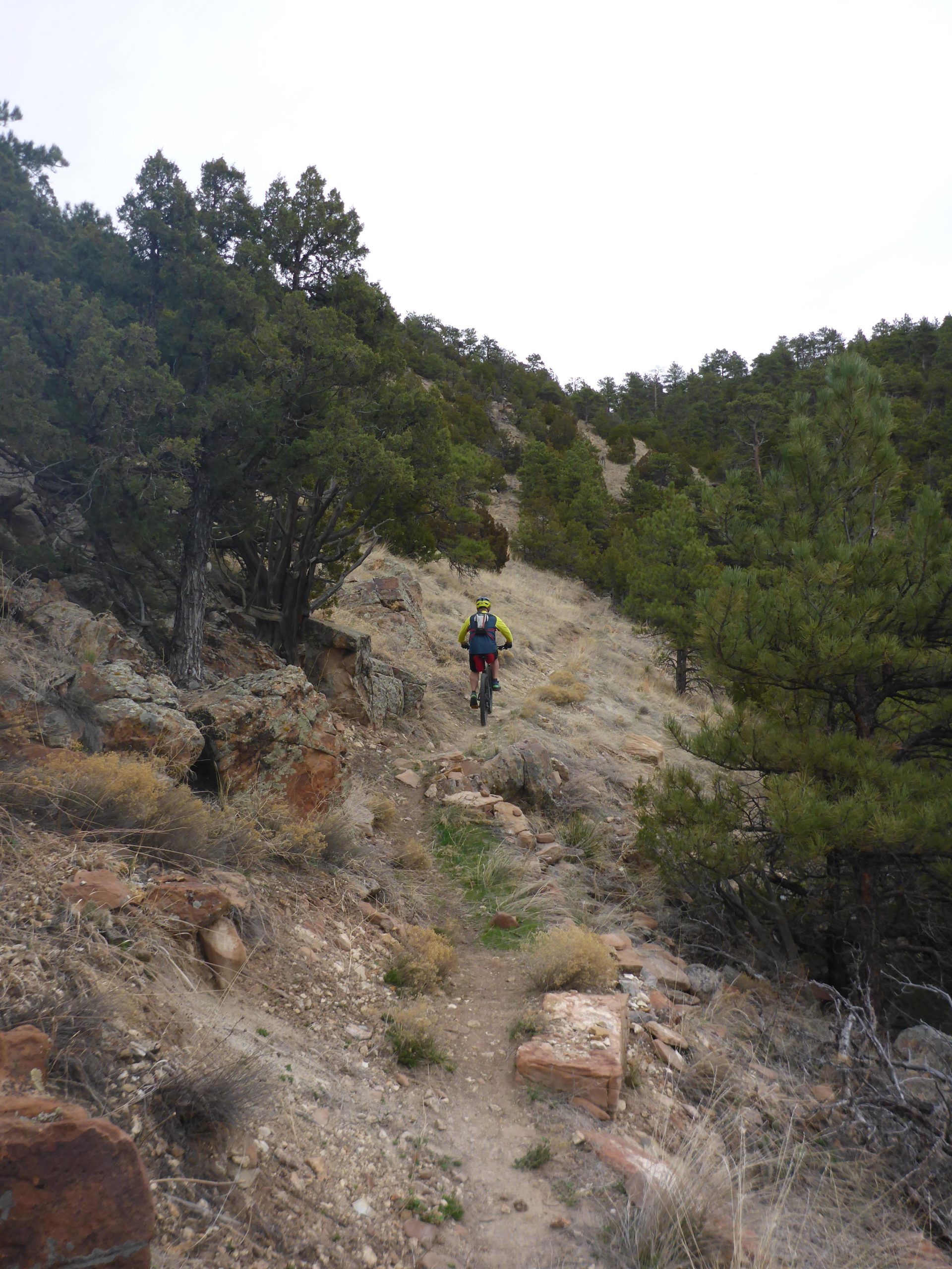 A hiker navigating a narrow, rocky trail surrounded by trees and vegetation on a hillside, with a cloudy sky above. Glendo State Park mountain bike trail.