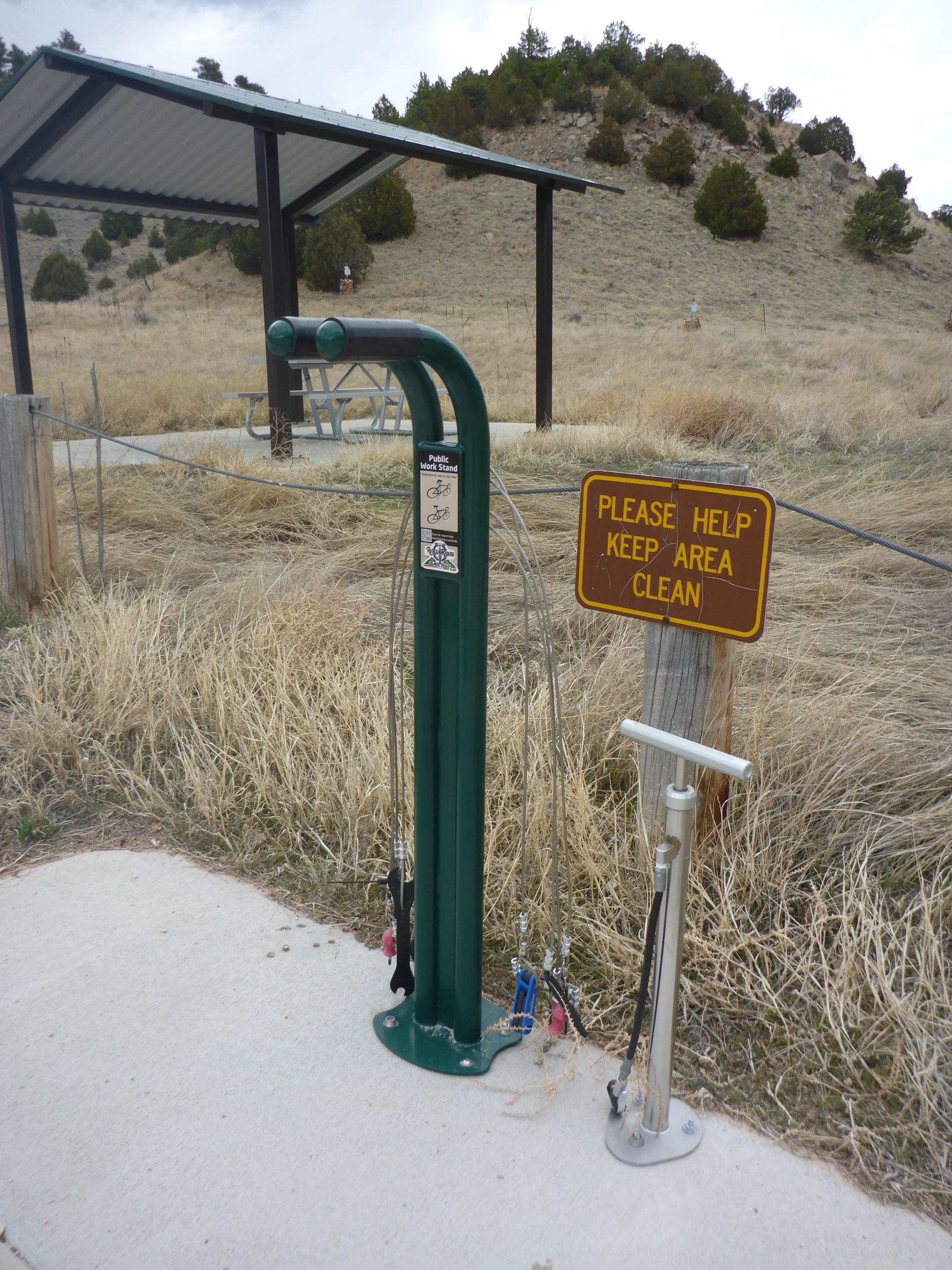 A public water station with a bicycle pump and maintenance tools, located next to a picnic area under a shelter. A sign nearby reads, "Please Help Keep Area Clean." The surrounding landscape features dry grass and low shrubs against a hilly backdrop. Glendo State Park mountain bike trail.