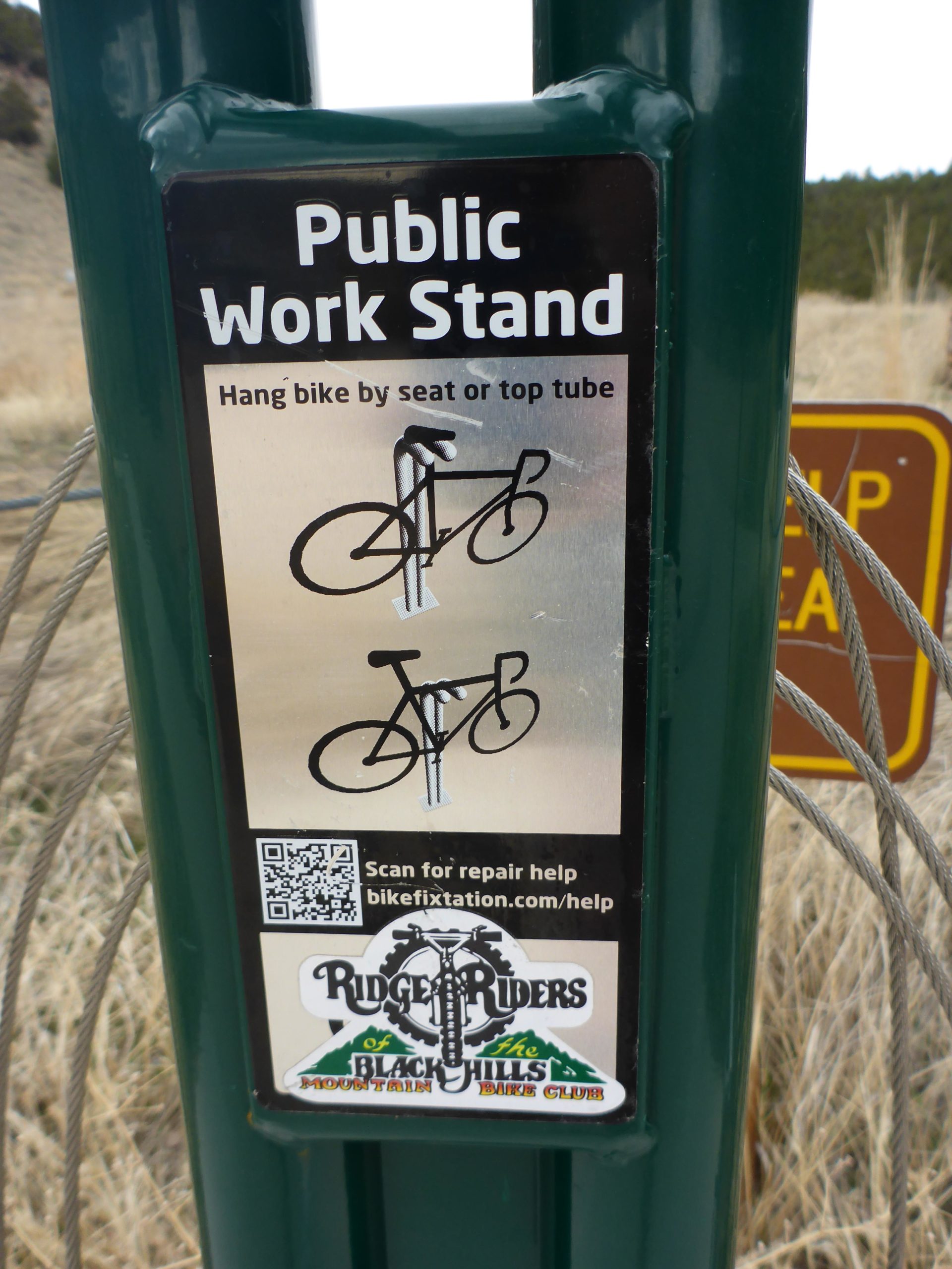 Public work stand for bicycles, featuring instructions to hang a bike by the seat or top tube. Includes a QR code for repair assistance and a logo for the Ridge Riders of the Black Hills mountain bike club. The background shows a natural landscape. Glendo State Park mountain bike trail.