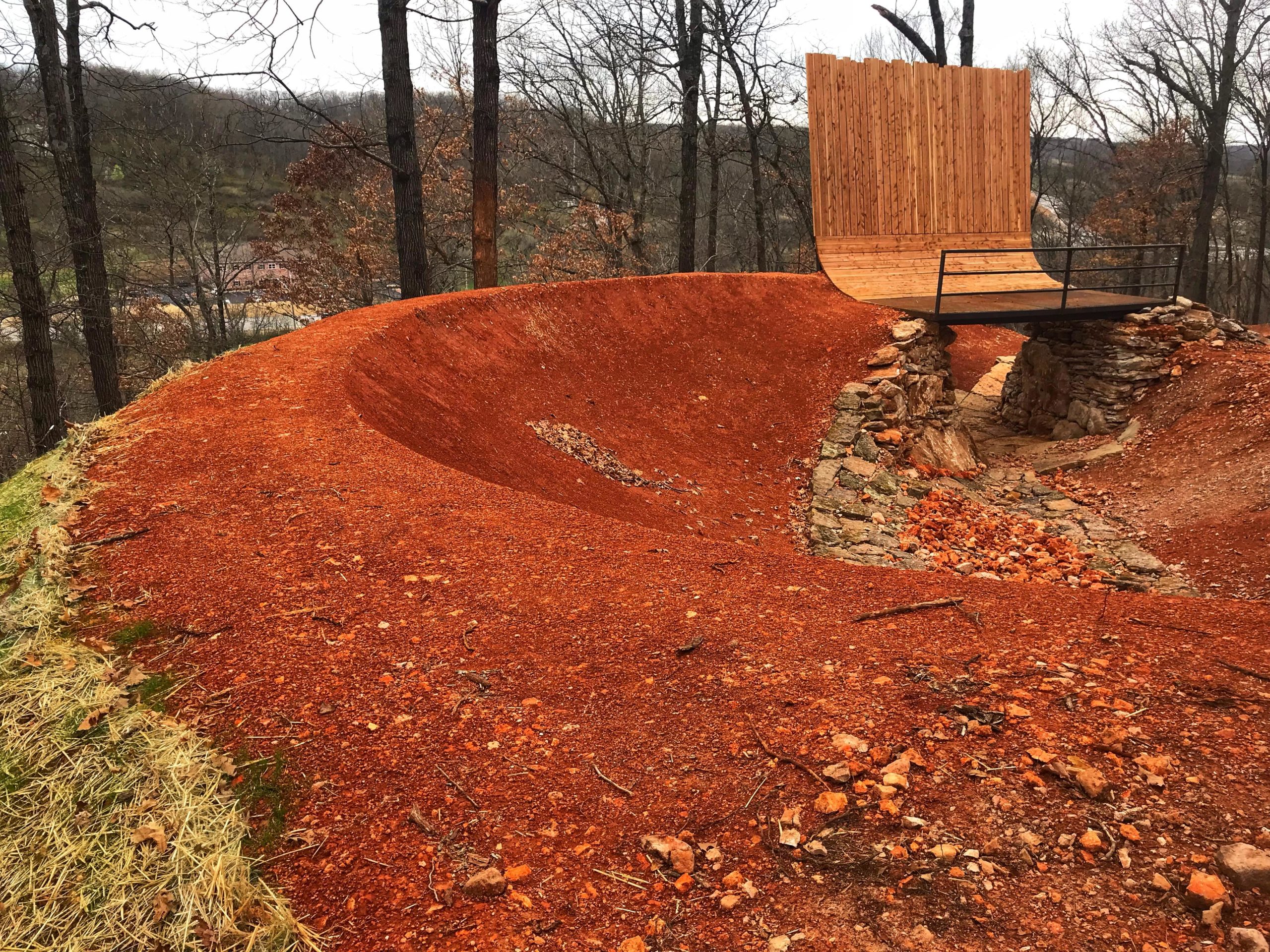 A dirt bike ramp and jump constructed from packed red clay, featuring a wooden wall ride at the far end. The area is surrounded by trees and rocky terrain, with scattered rocks and stones visible on the ground. Slaughter Pen Trail mountain bike trail.